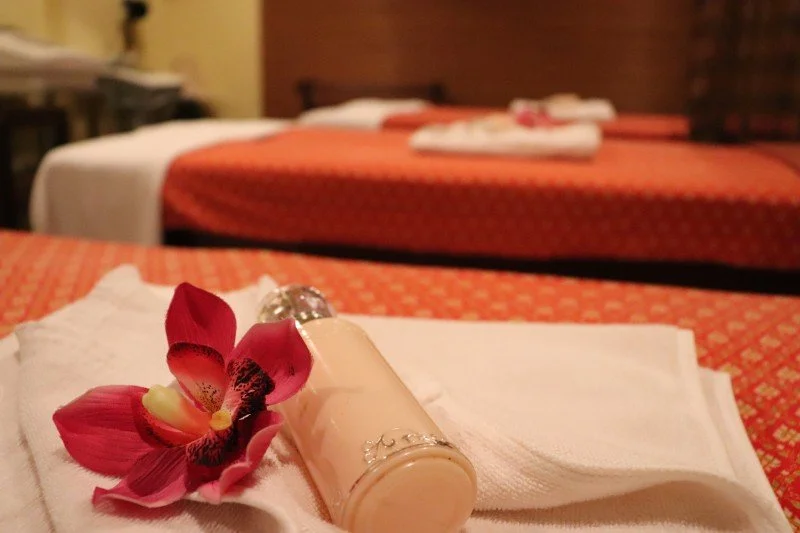 A hotel bed with folded towels, a small bottle, and a pink flower on a white cloth in the foreground.