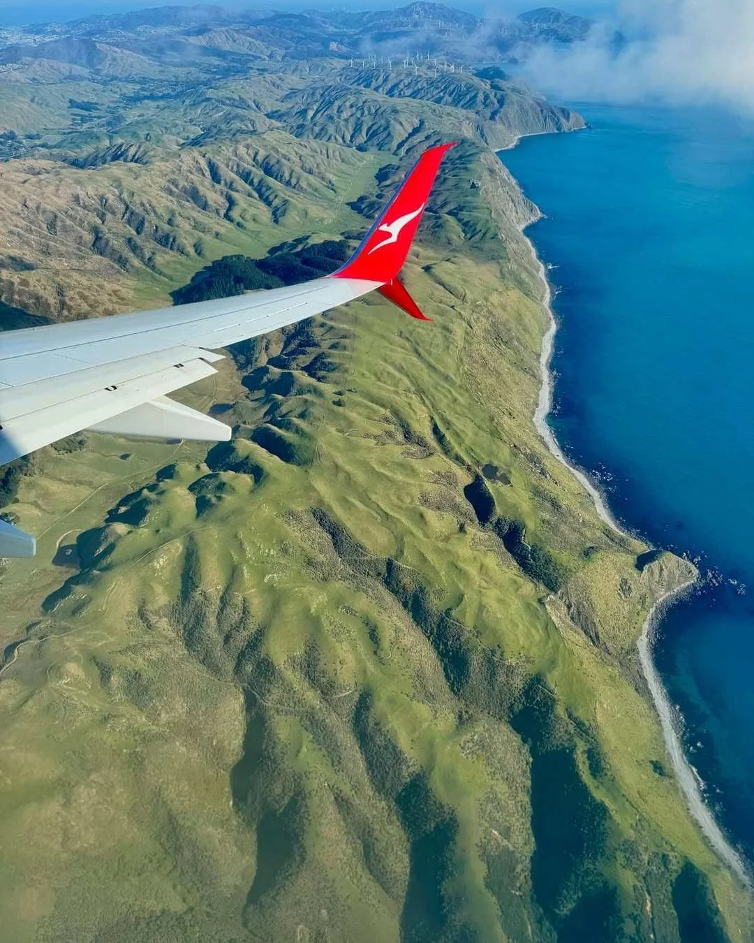 Aerial photo of the New Zealand coastline taken from a Qantas plane window, showing green hills meeting bright blue ocean.