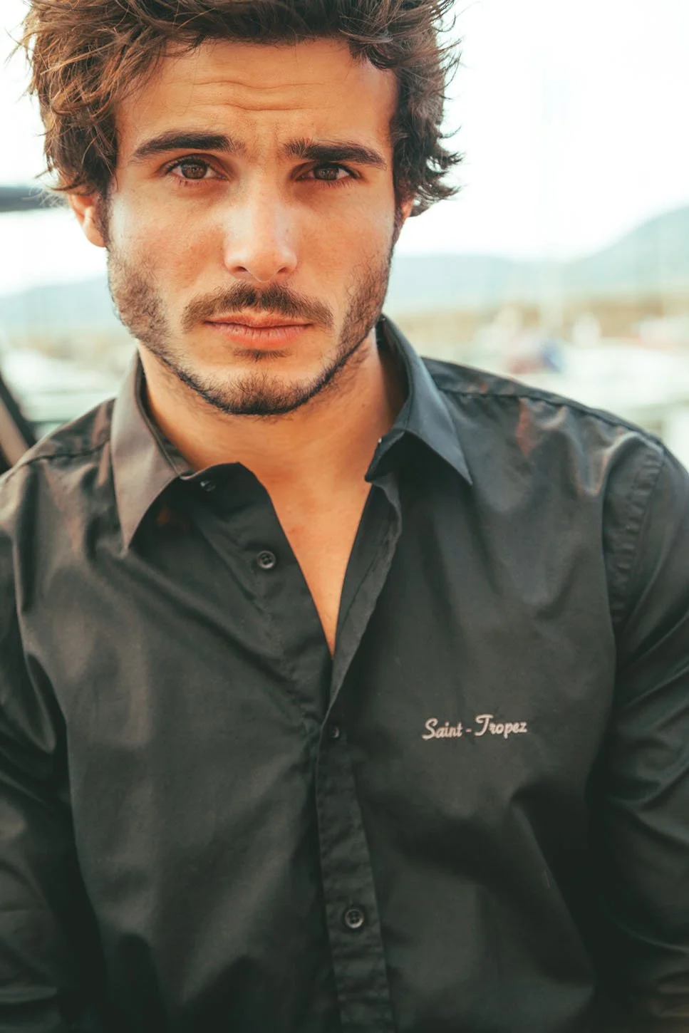Jeune homme aux cheveux bruns, barbe légère, portant une chemise noire avec inscription "Saint-Tropez", regardant vers l'objectif avec un fond flou de paysage extérieur.