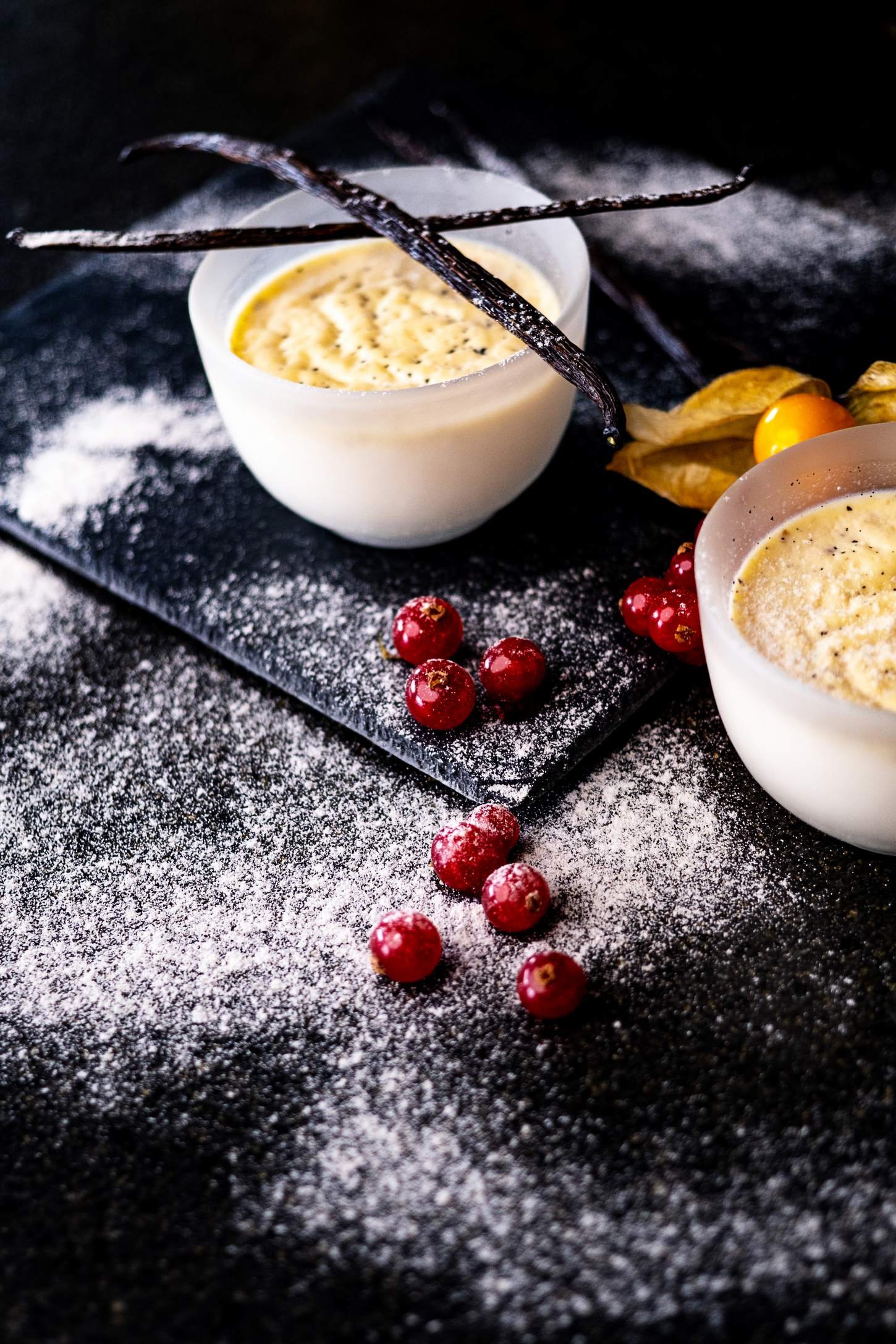 Deux bols de crème dessert avec des garnitures, entourés de groseilles rouges, sur une plaque noire avec de la farine ou du sucre poudré, décorée de branches d'anis étoilé et d'une physalis.