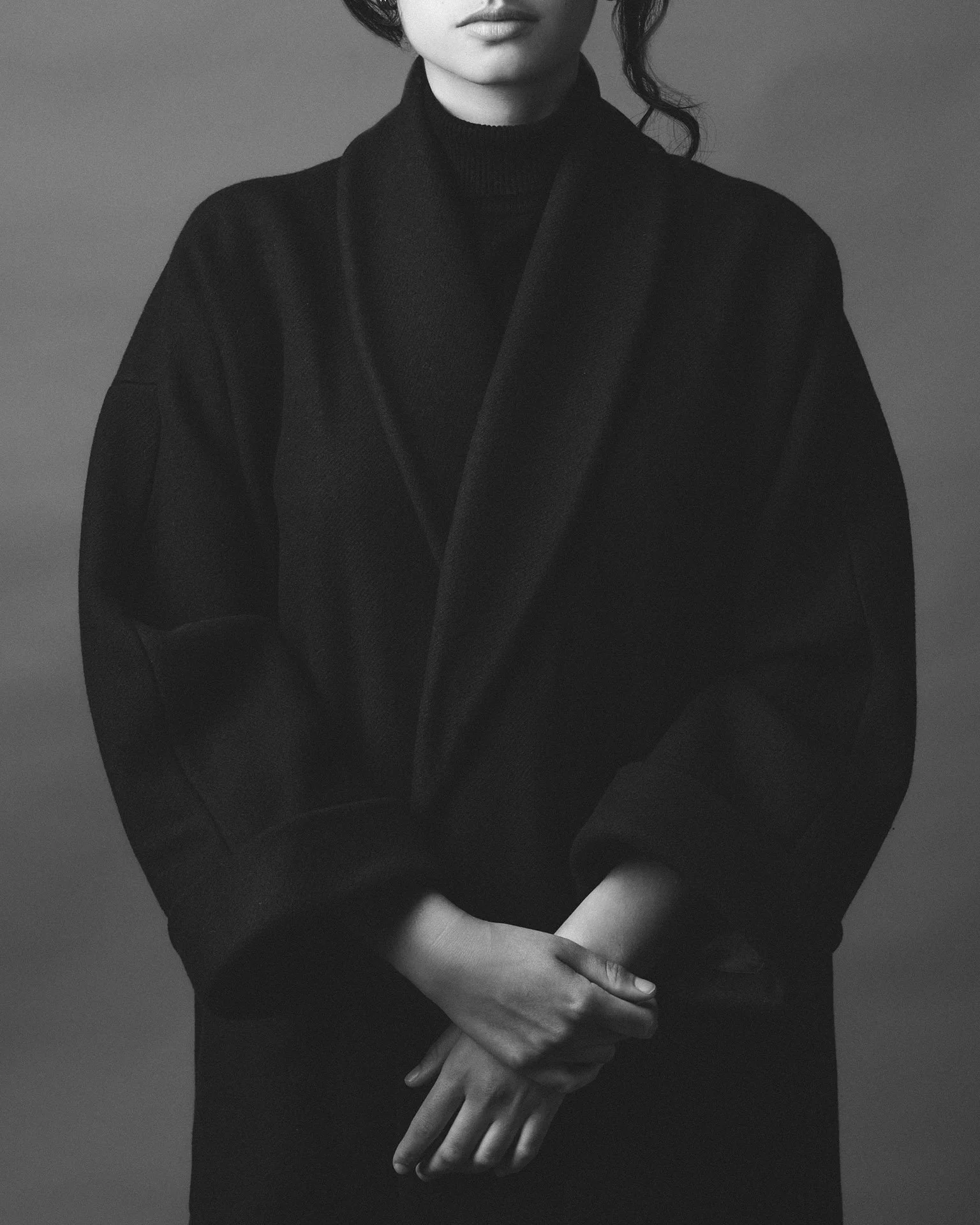 Une femme en noir et blanc, portant un manteau et un turtleneck, avec les mains croisées devant.