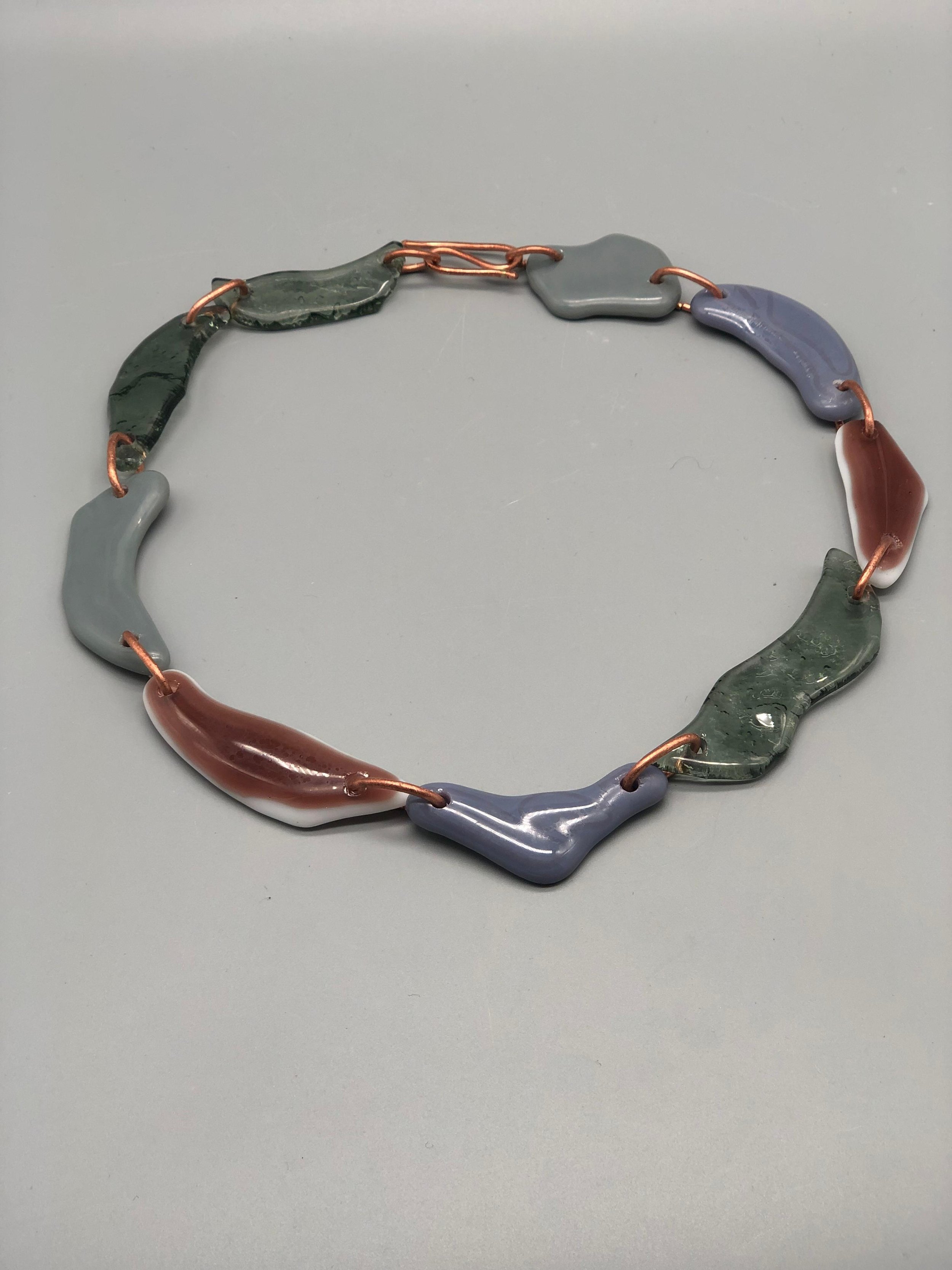 Manumara Arte Design — contemporary necklace in fused glass with organic links in green, grey, lilac and terracotta tones, available at Crepundia Gioielli Bologna.