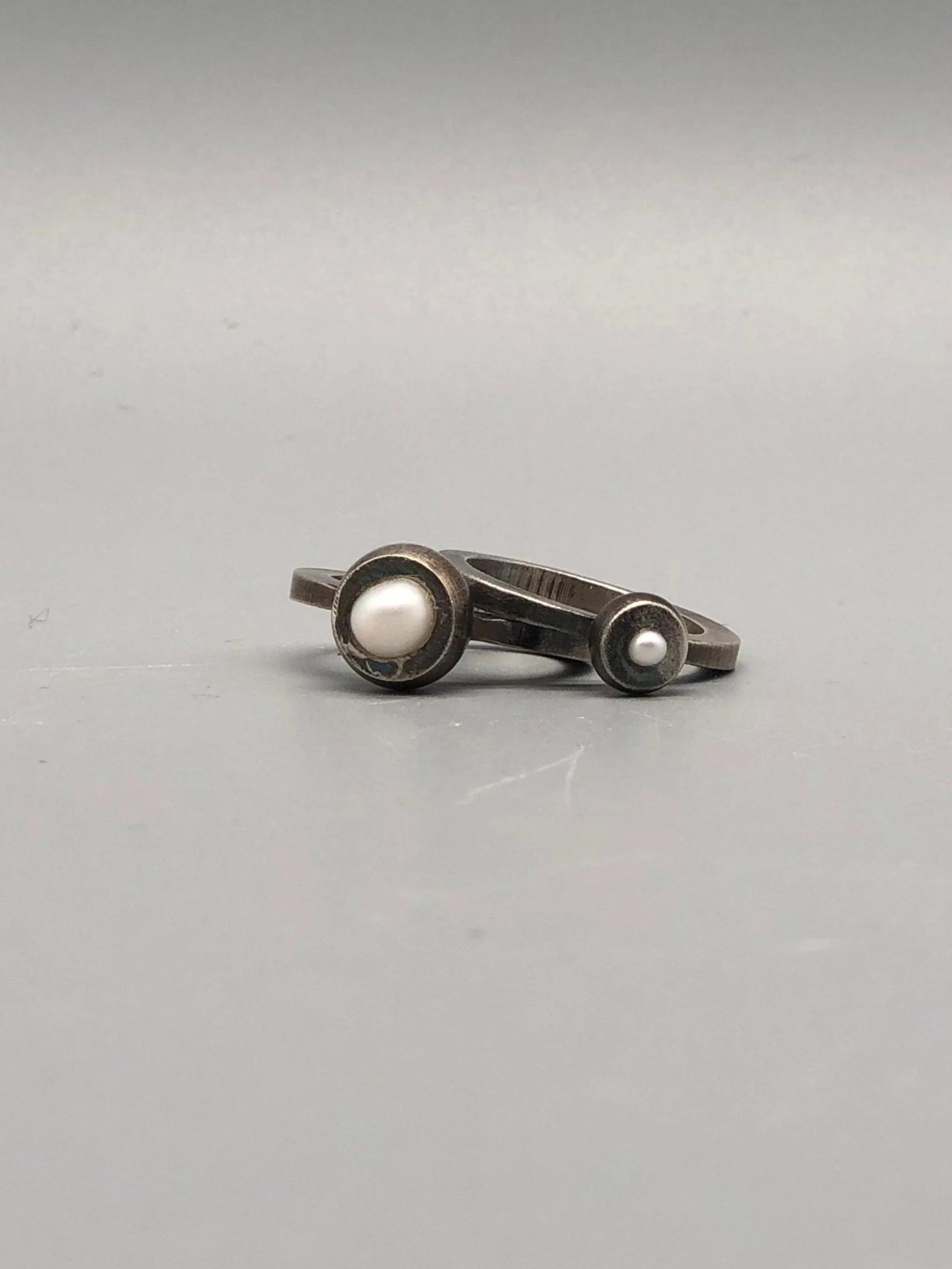 Anelli Bulls mini e maxi con perla giapponese.
Argento 925 brunito, forme essenziali e dettagli discreti reinterpretano la perla in chiave contemporanea.

Bulls mini and maxi rings with Japanese pearl.
Oxidized sterling silver, essential forms, and d