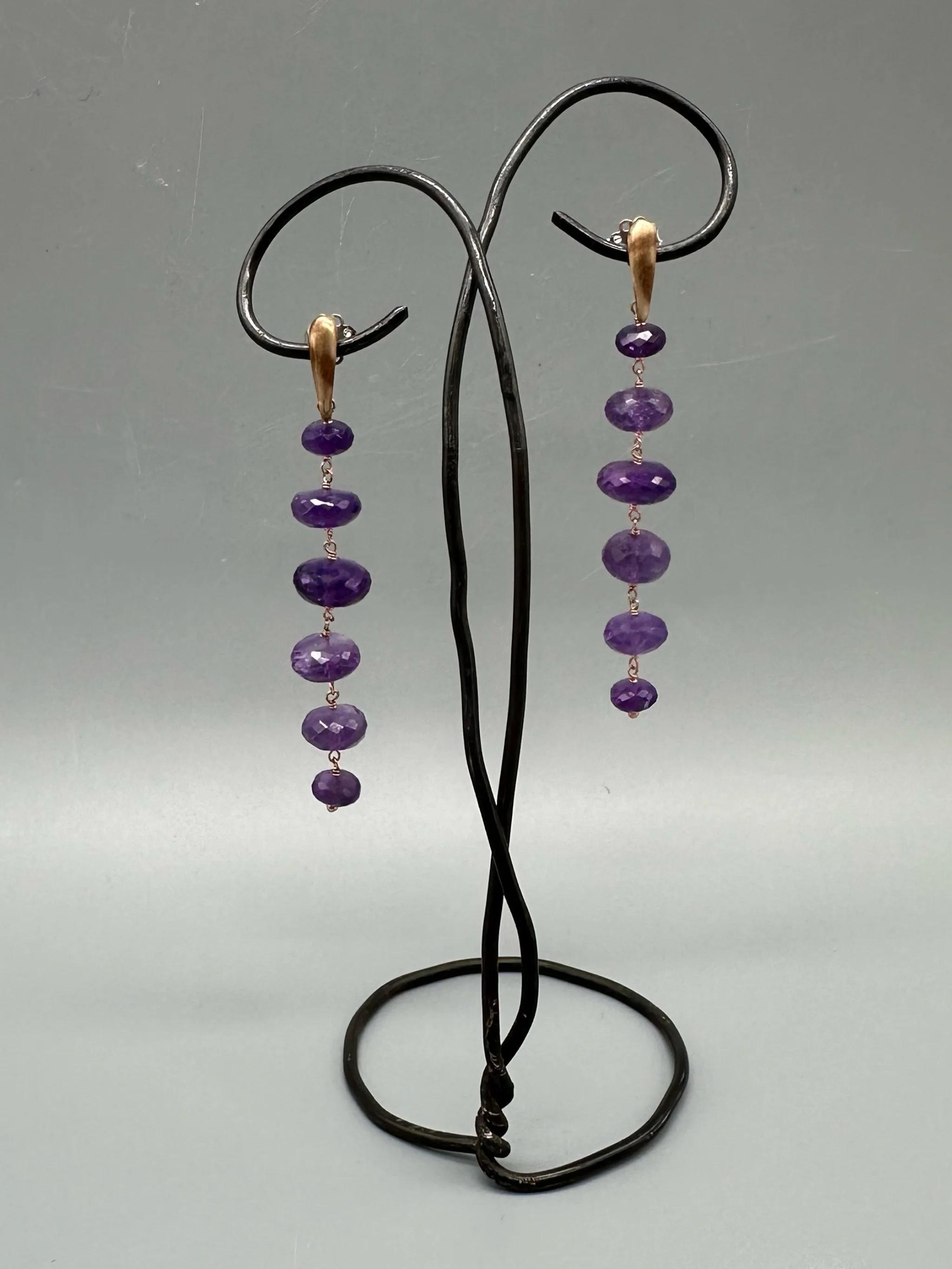 Orecchini Bold Ametista con pietre viola sfaccettate e montatura essenziale. Bold Amethyst earrings with faceted purple gemstones and a minimal metal setting.