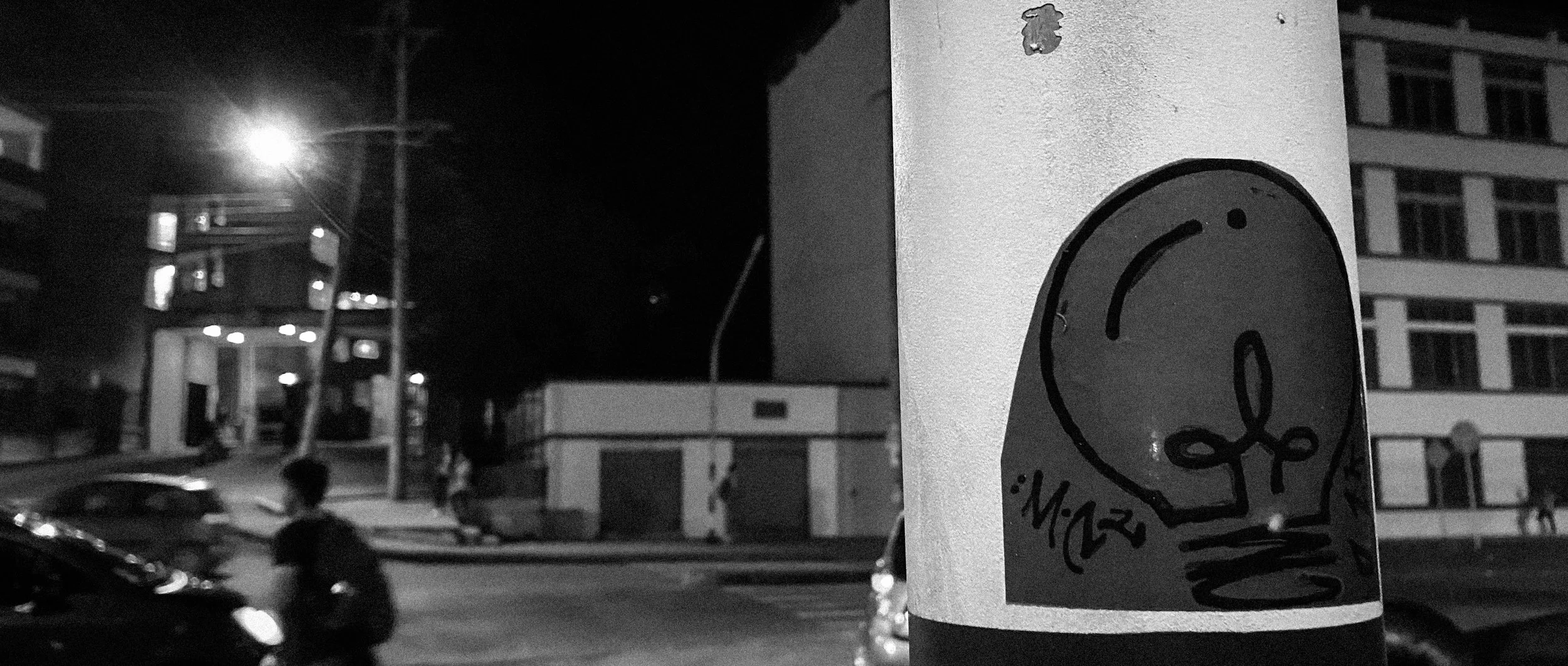 Street scene at night with a sticker of a smiling face on a pole in the foreground, blurred cars and buildings in the background, and streetlights illuminating the scene.
