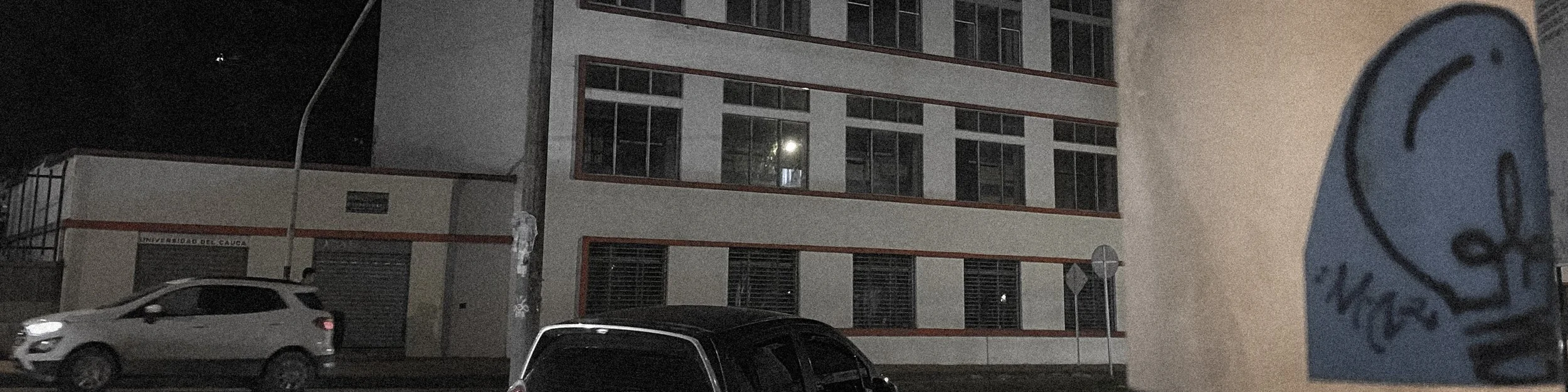Nighttime street view with parked cars and a building with lit windows.