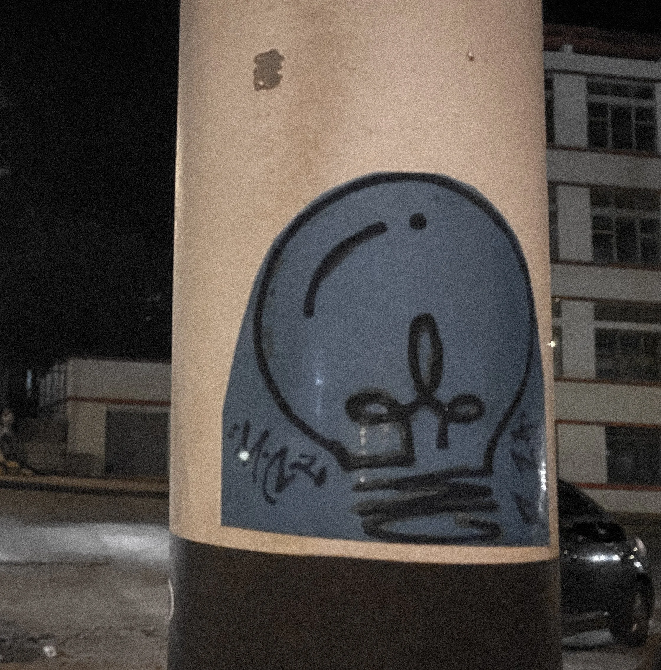 Street art graffiti of a cartoonish light bulb with a smiling face and a bow tie, painted on a beige utility box at night.