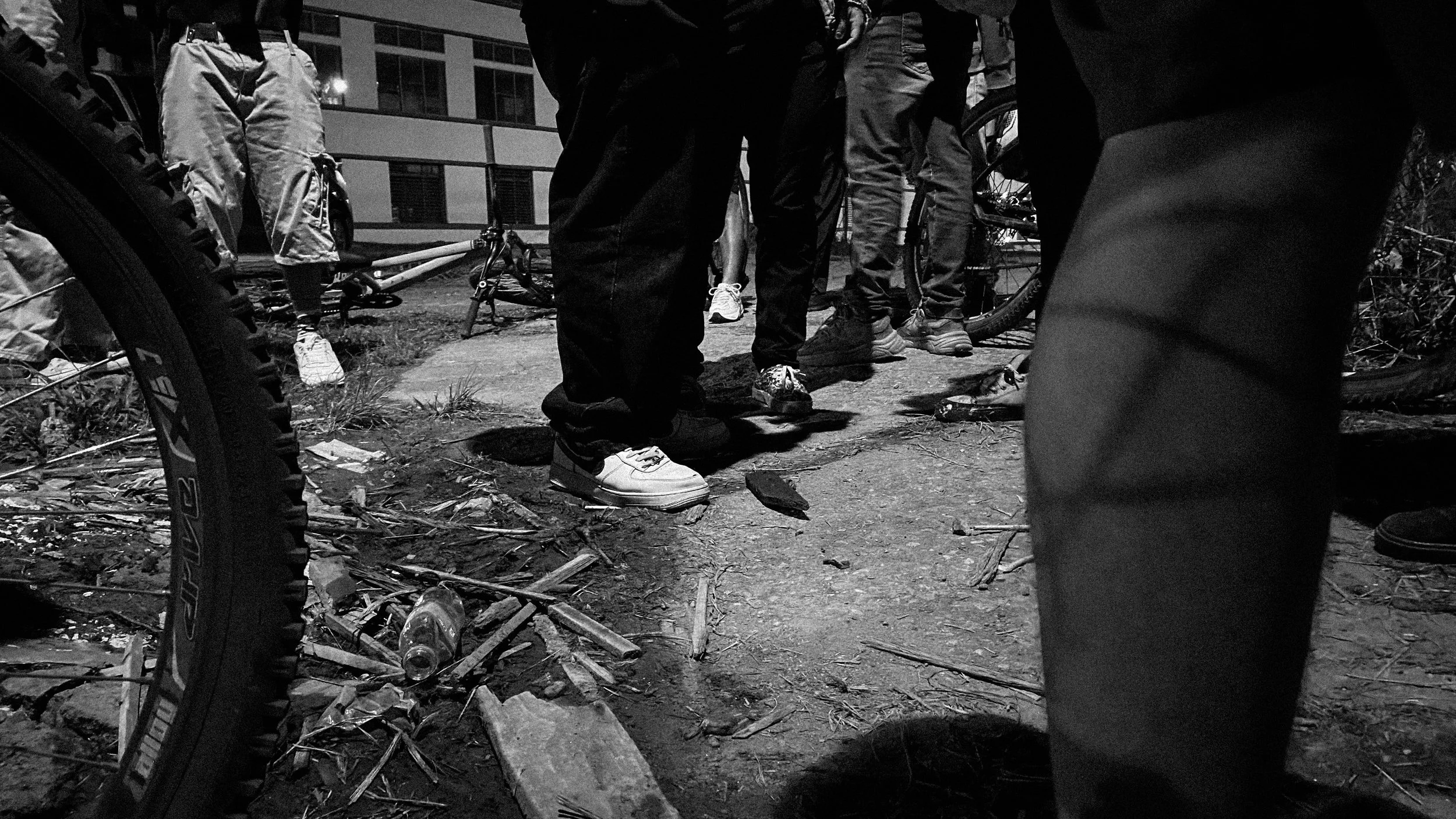 People standing outdoors on a sidewalk at night, captured from a low angle, with bicycles and debris on the ground.