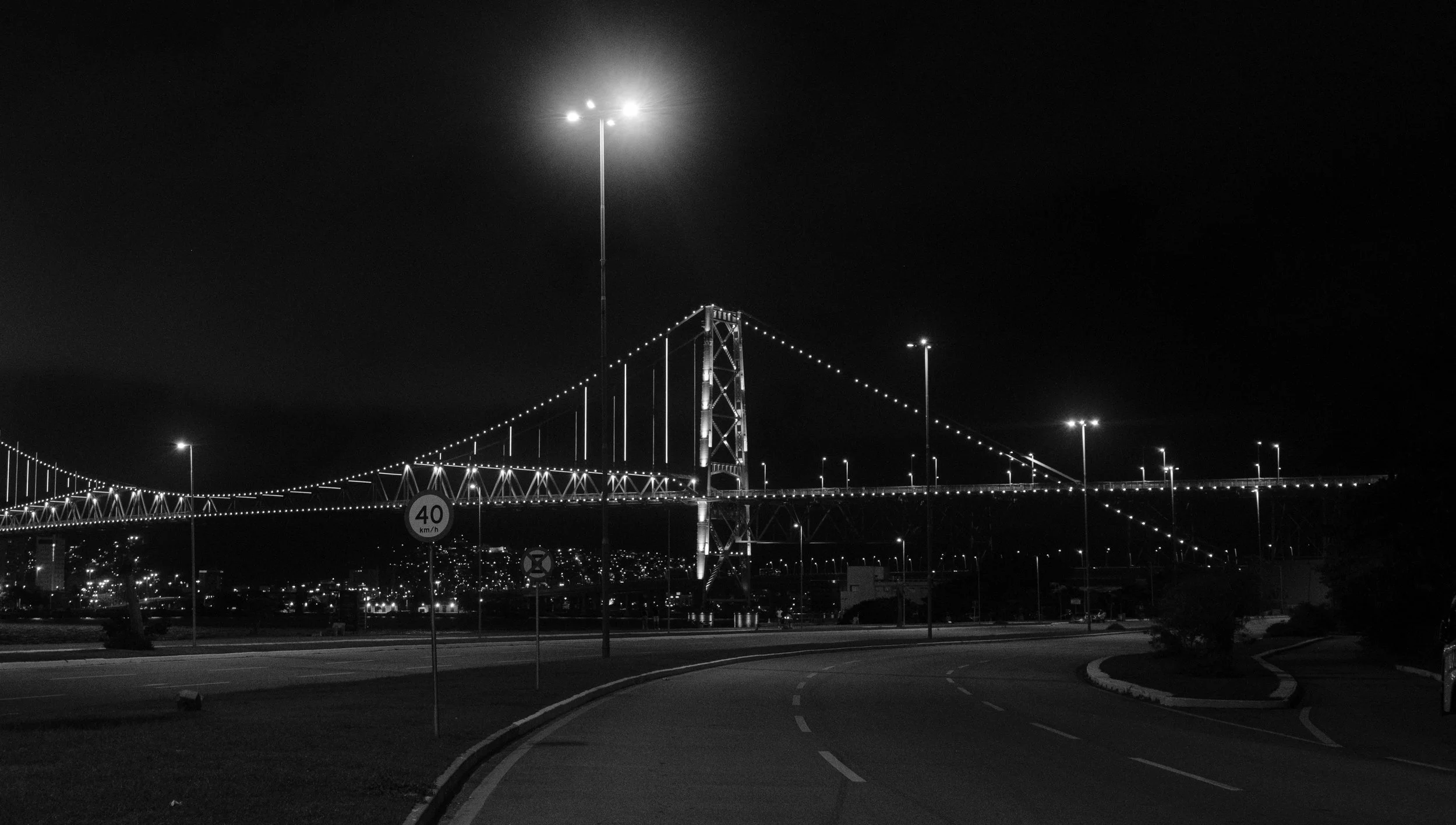 Night view of a lit suspension bridge spanning a body of water with city lights in the background.