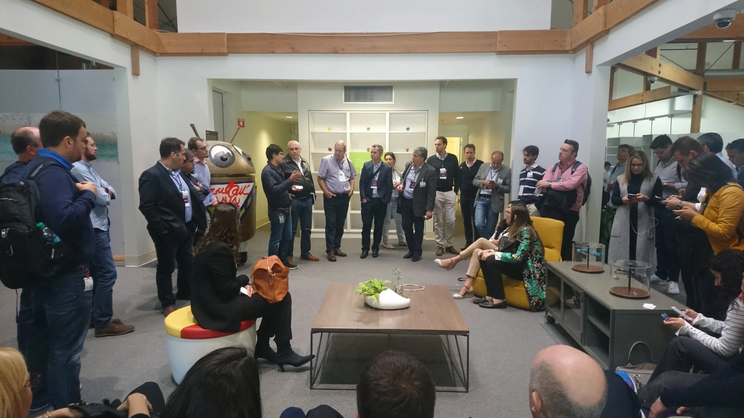 Group of people gathered in a circular formation in an indoor space, with some sitting on chairs and others standing, listening to a woman speaking. The setting appears to be an office or conference area, with a decorative table, a yellow armchair, a