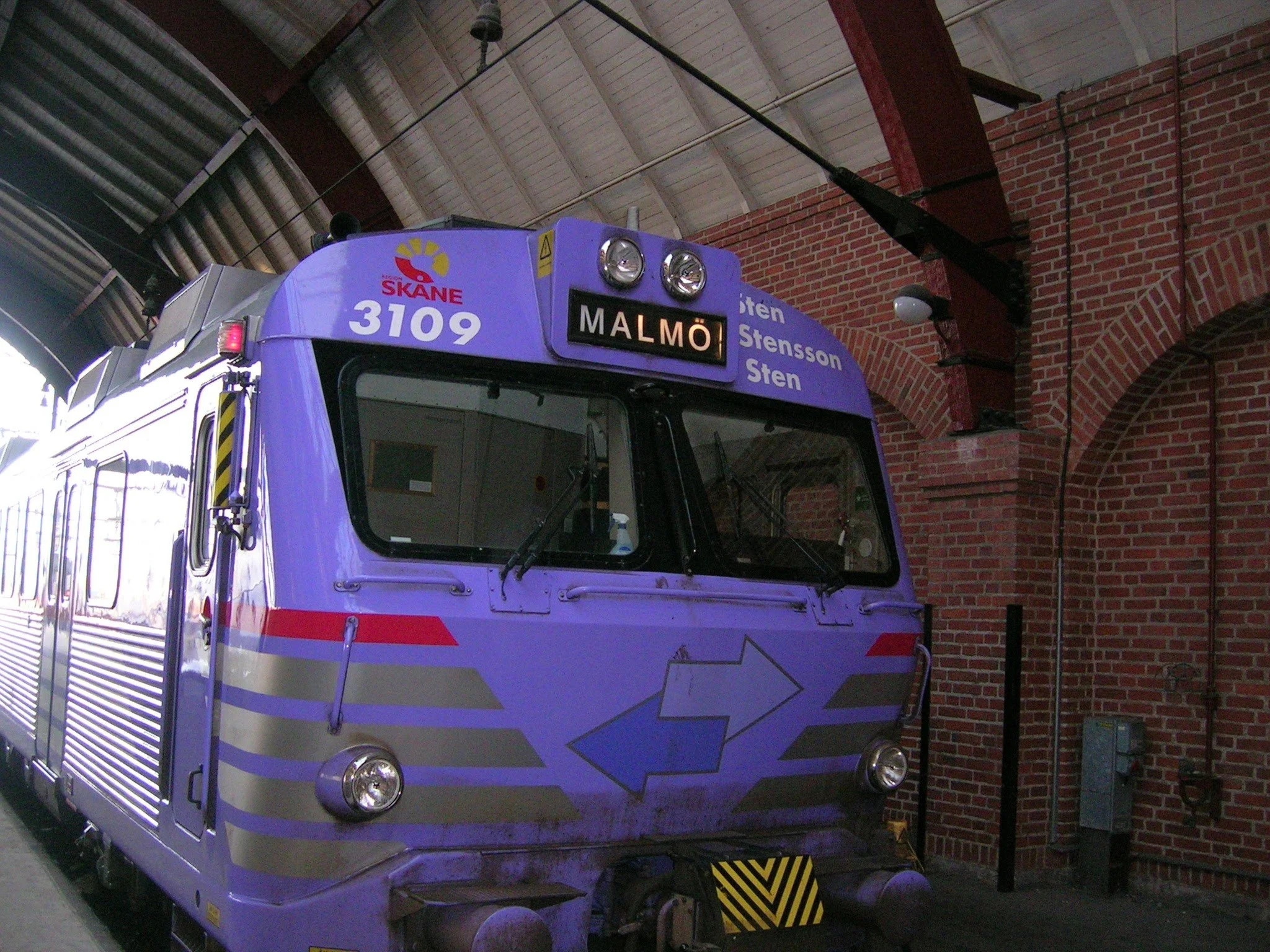 A purple train at a station platform with signs indicating 'Malmö' and route numbers, inside a brick train station with a high arched ceiling.