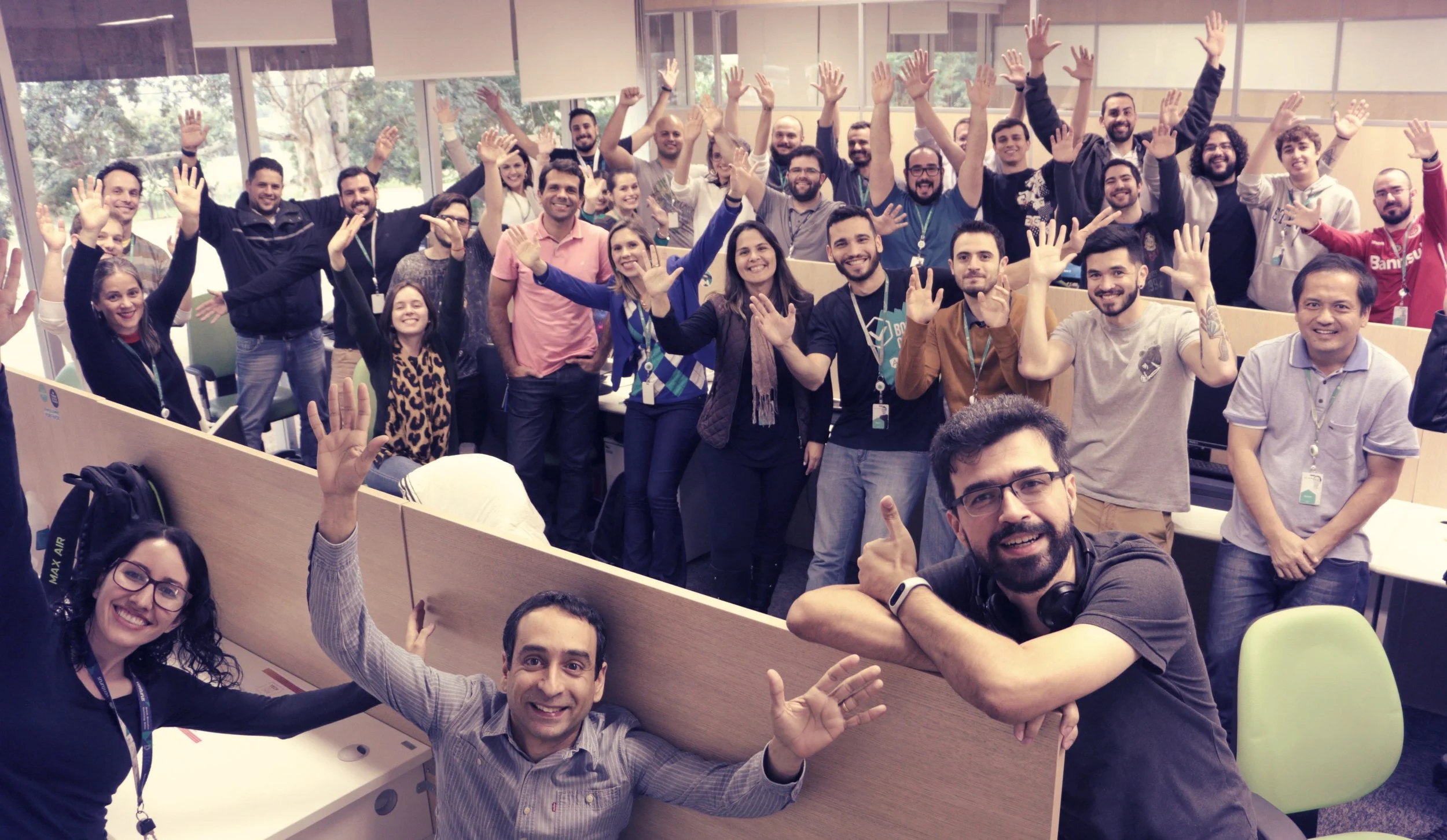 Group of coworkers in an office celebrating, smiling, and waving at the camera, with some sitting and others standing.