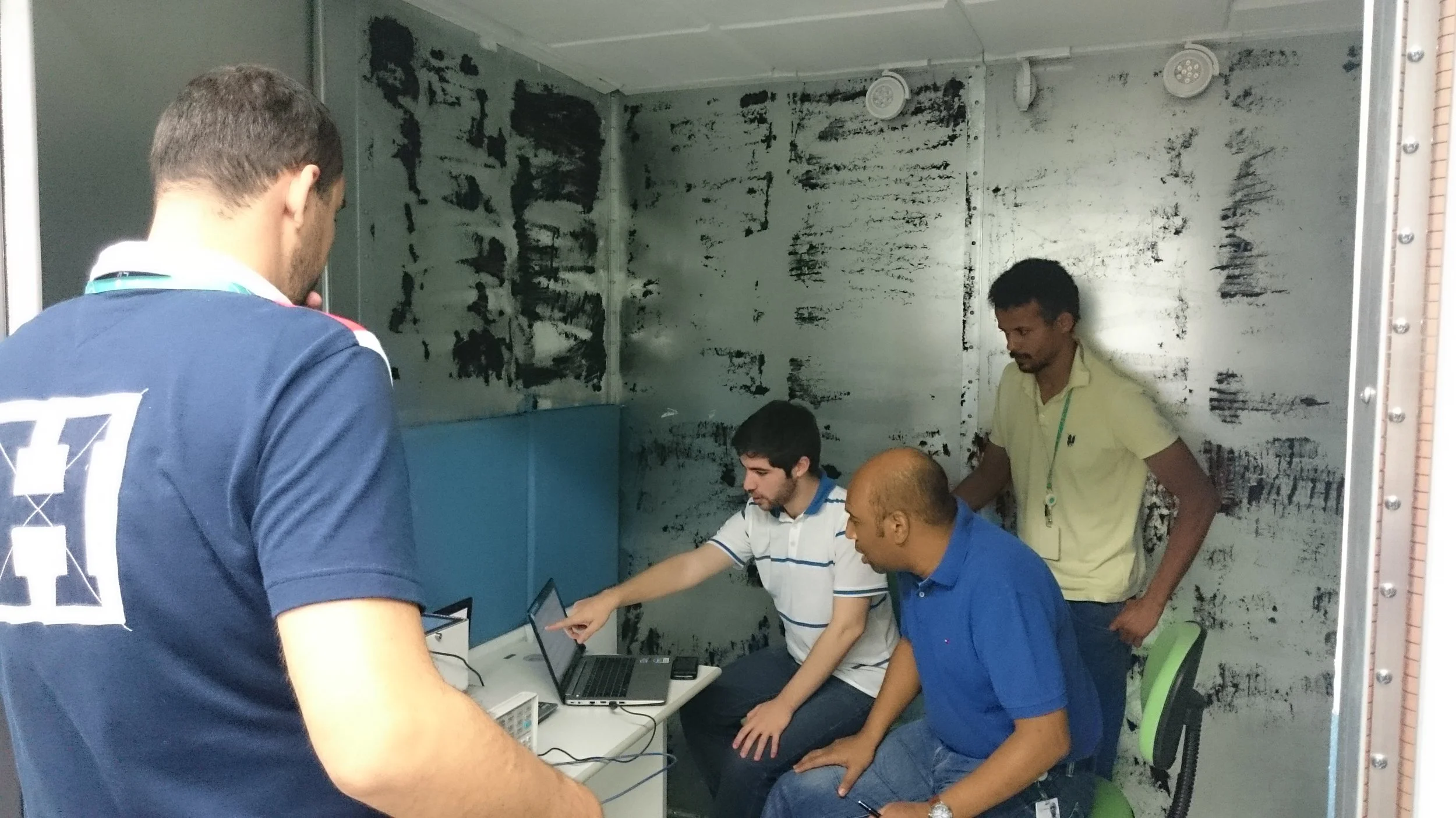 Four men inside a small room with metallic walls, two are seated at a table with laptops, one is standing and pointing at a laptop screen, and the fourth man is standing, observing.
