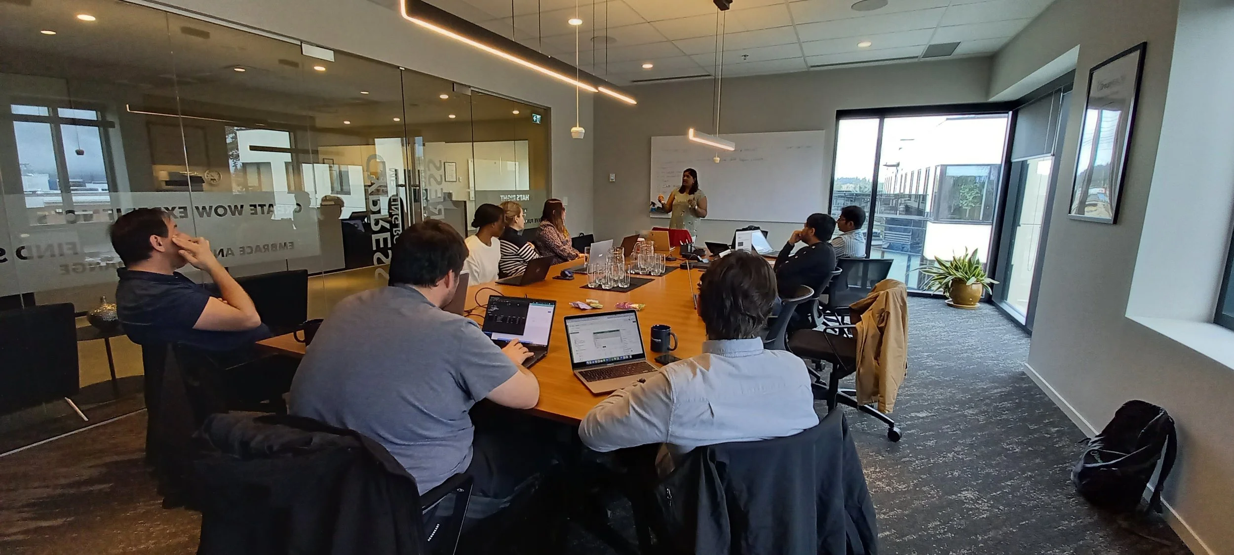 A business meeting or presentation taking place in a modern conference room with multiple attendees seated around a large oval table, some working on laptops, listening to a woman standing and speaking at the whiteboard. The room has large windows le