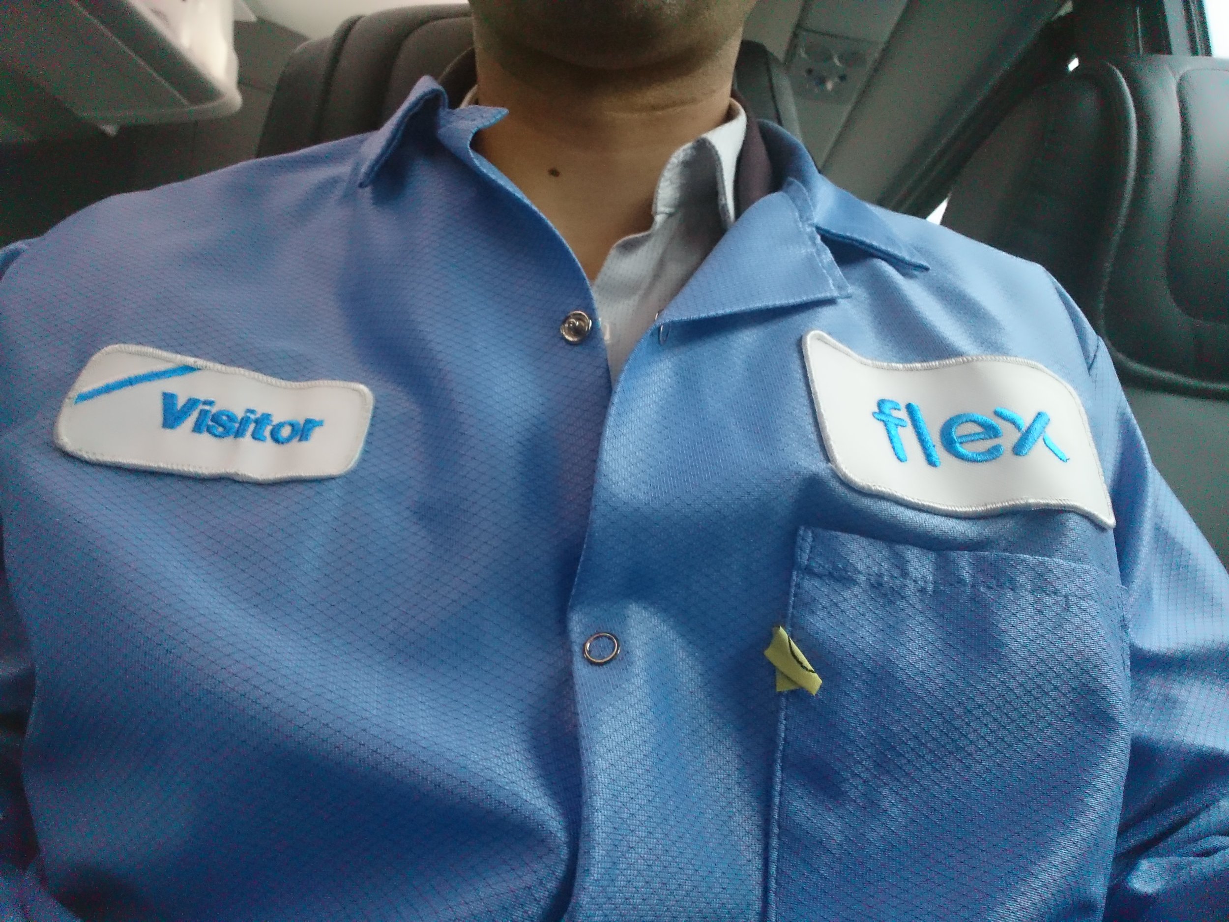 Close-up of a person wearing a blue uniform with 'Visitor' and 'flex' patches, seated inside a vehicle.