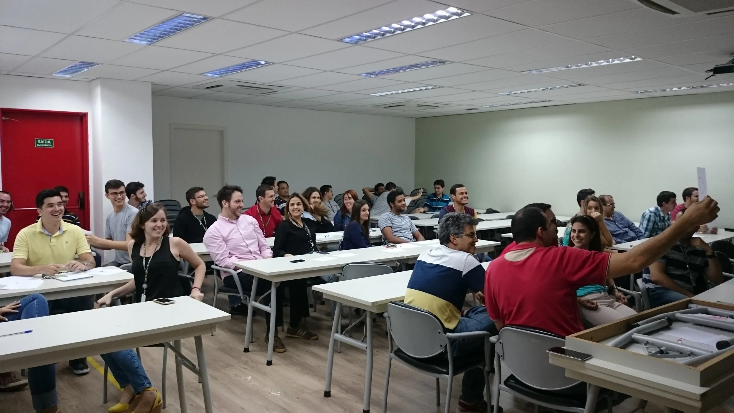 A group of people sitting in a classroom or conference room, smiling and paying attention to a person taking a selfie at the front. The room has white walls, a red door, and fluorescent ceiling lights.
