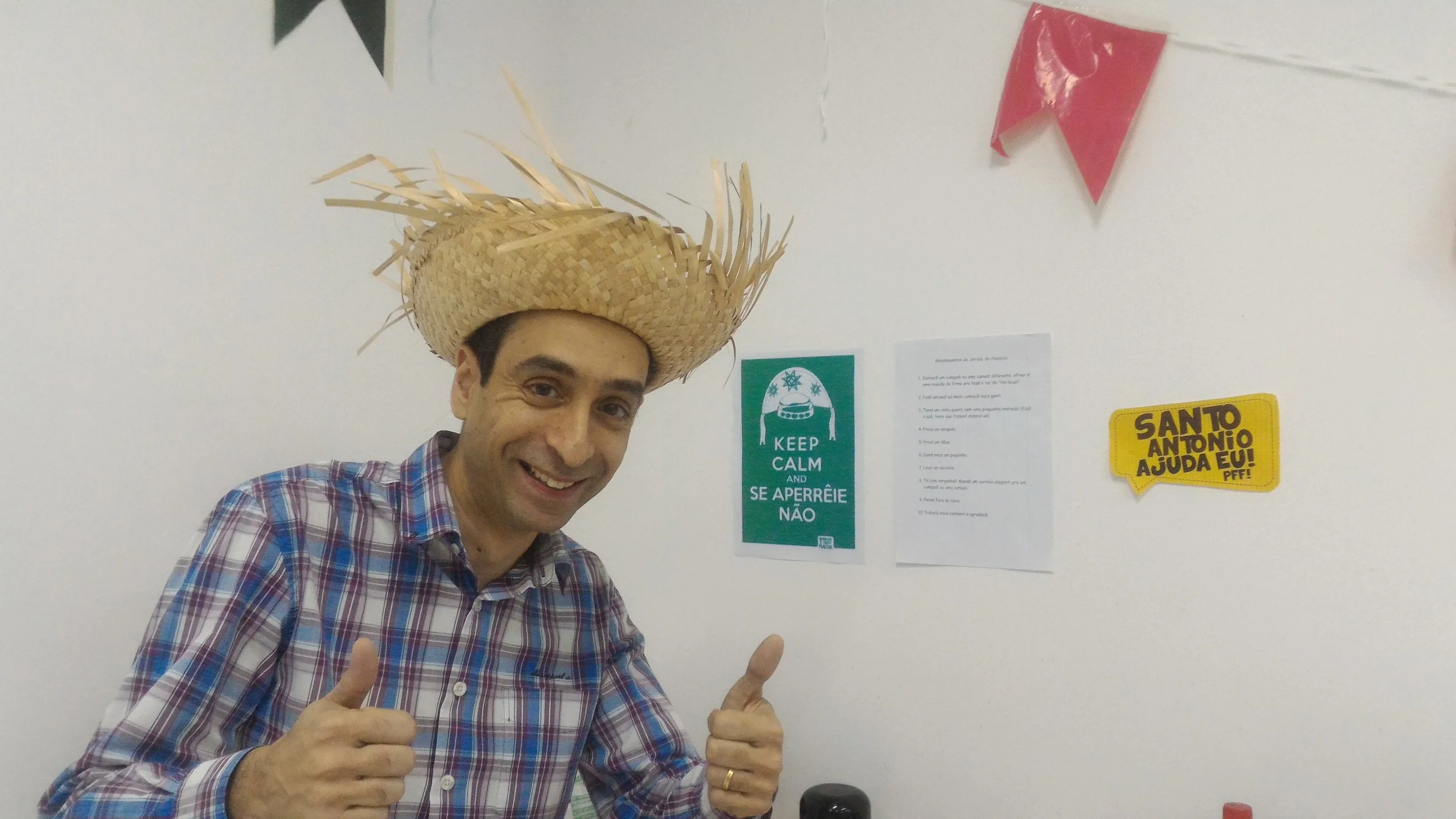 A man wearing a plaid shirt and a straw hat is giving two thumbs up and smiling, standing indoors against a white wall with various posters and decorations.