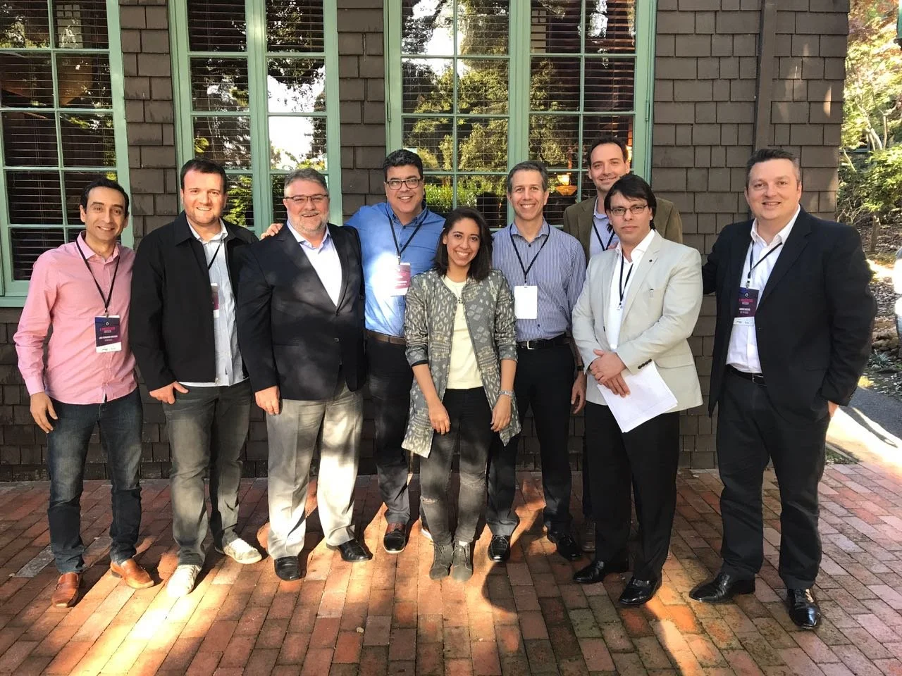 A group of ten diverse professionals standing together outdoors in front of a brown brick building with green-framed windows. They are smiling and dressed in business casual to formal attire, with some wearing conference badges around their necks.