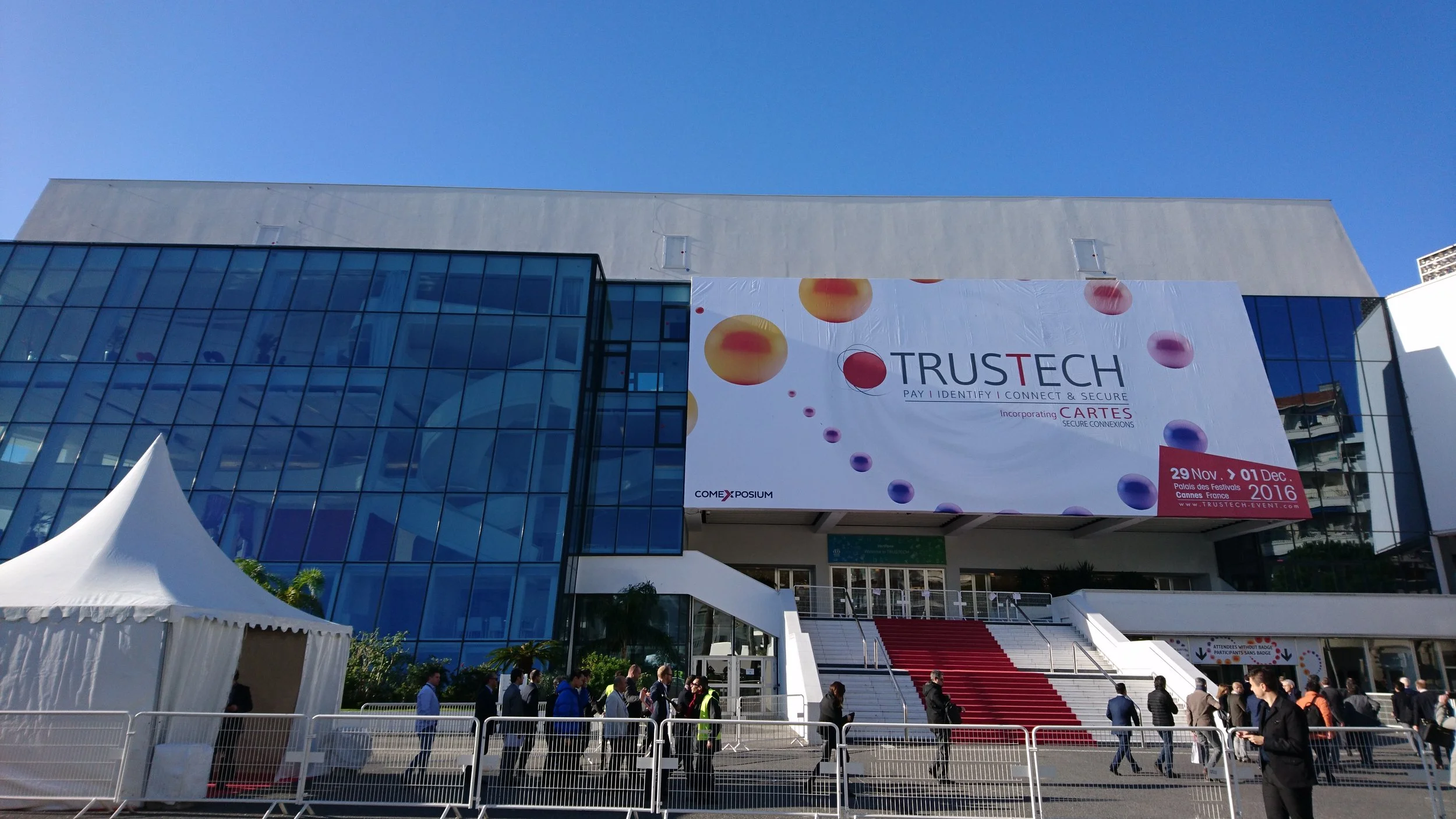 People walking into a convention center with a large banner for TRUSTECH event, displaying dates and branding, and a white tent outside.