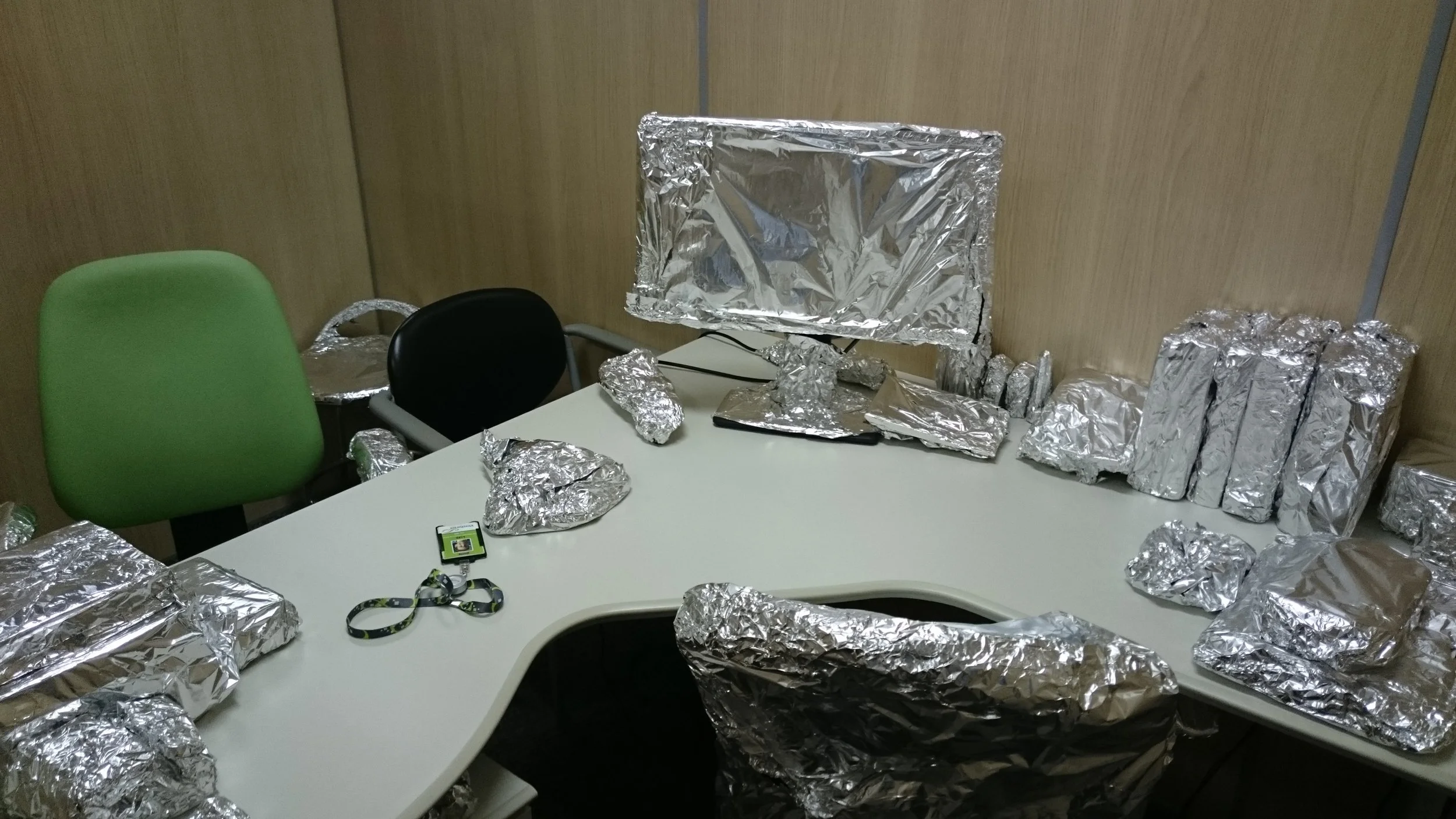 Office conference room with a desk covered in crumpled aluminum foil, a computer monitor also wrapped in foil, and chairs with foil on their seats.