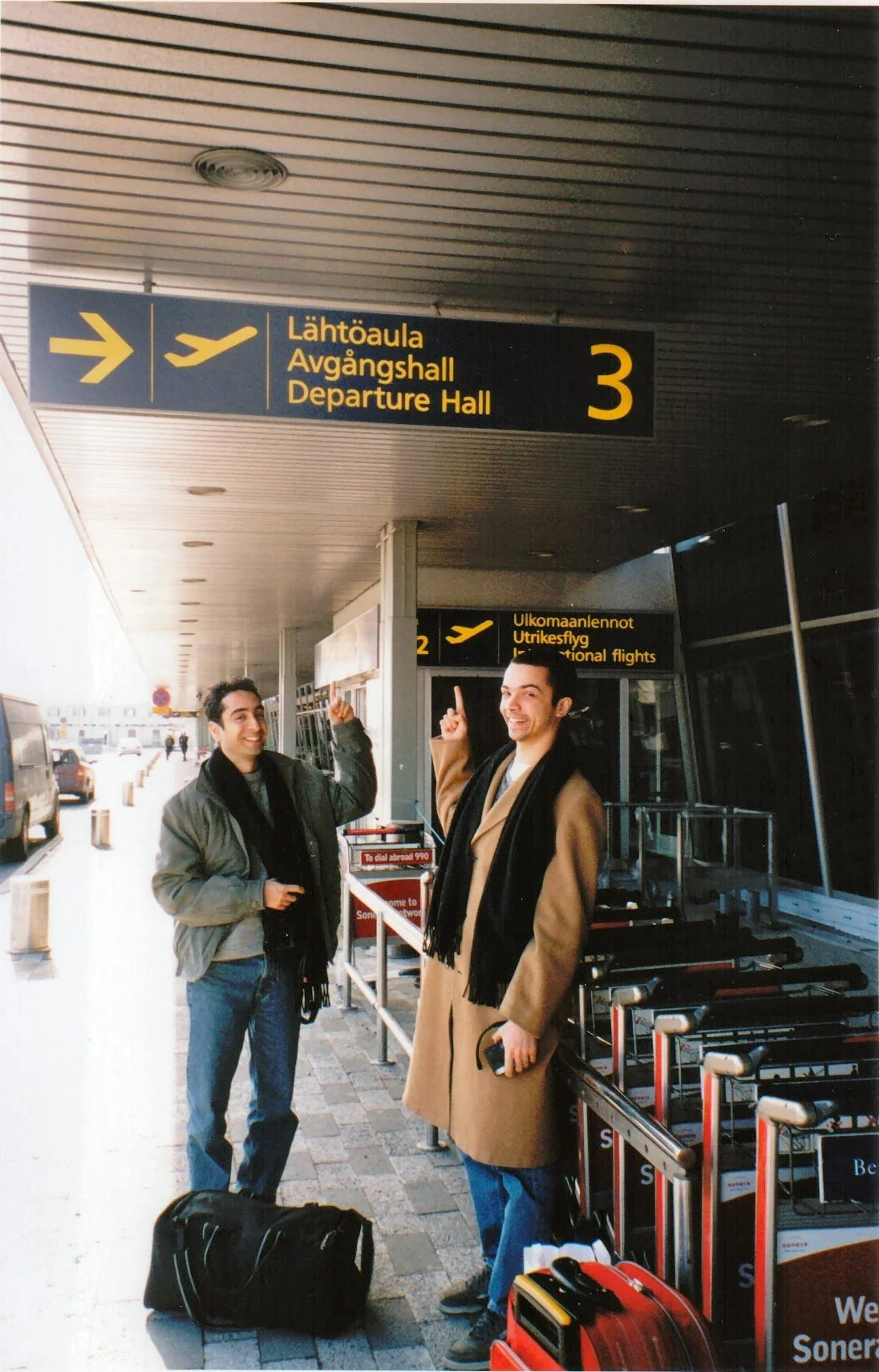 Two men standing outside a departure hall at an airport, smiling and pointing upwards, with luggage on the ground nearby.