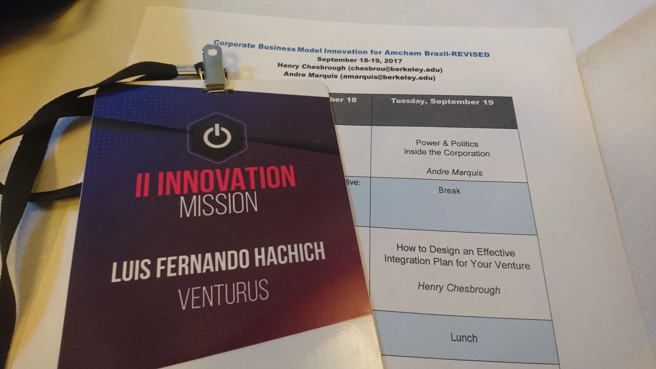 Conference badge and schedule sheet on a table. The badge reads 'Innovation Mission' with the name Luis Fernando Hachich from Venturus, and the schedule lists events including a talk by Andre Marquis and a session on designing integration plans.
