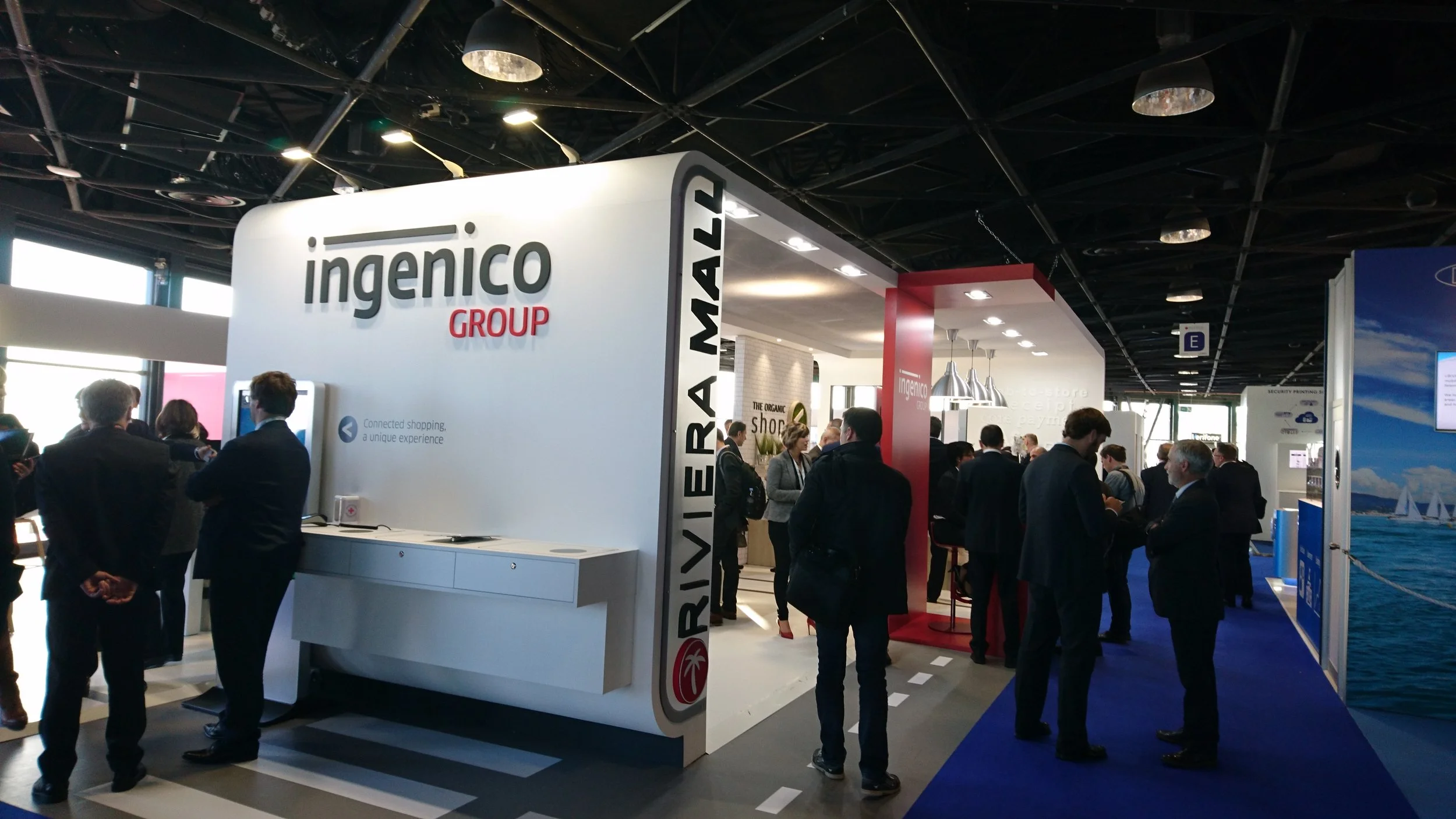 Trade show booth for Ingenico Group with people conversing and exploring exhibits, with a blue carpeted walkway and modern lighting.