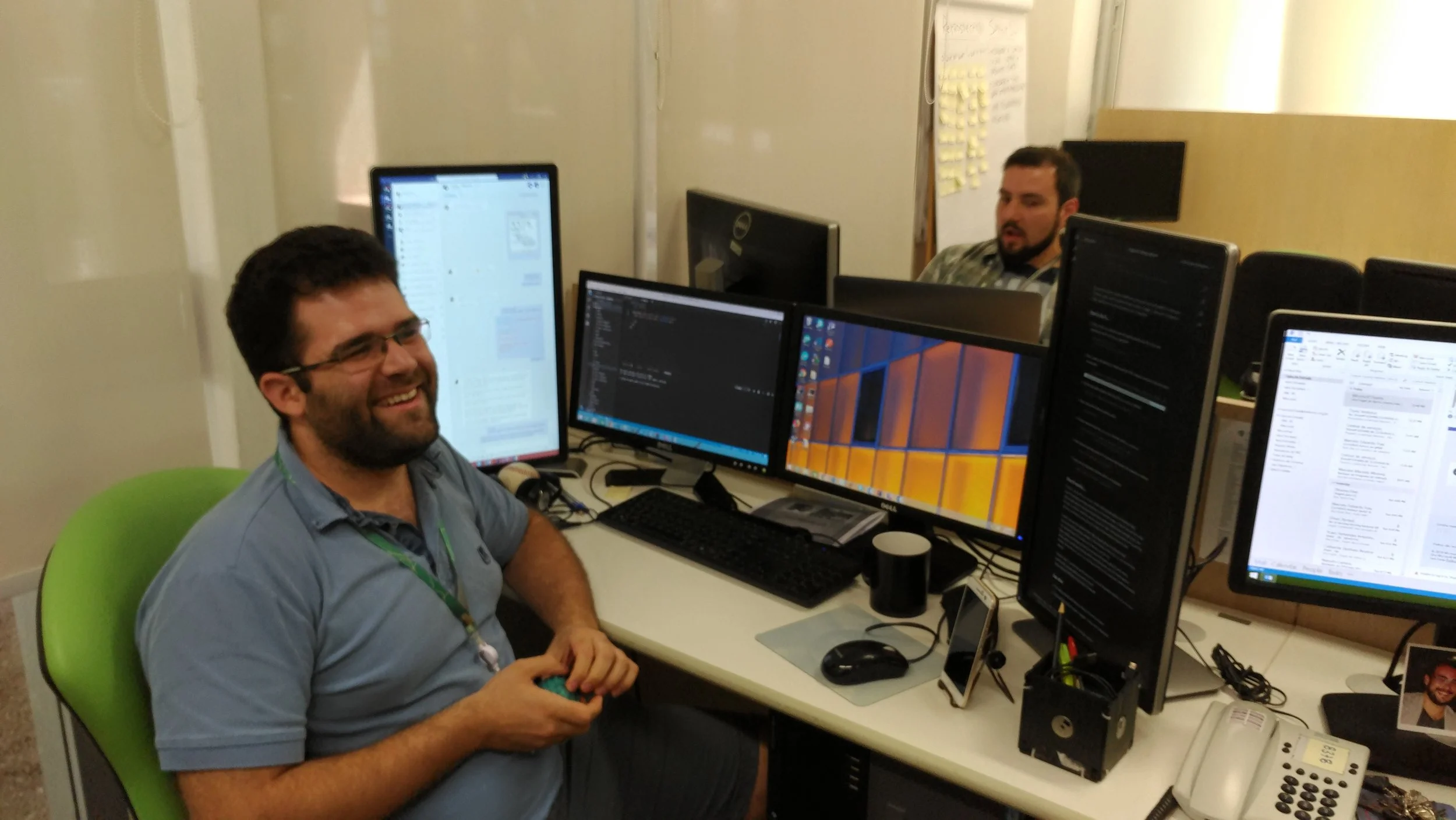 Two men working at a shared desk with multiple monitors, one smiling and the other focused, in an office setting.