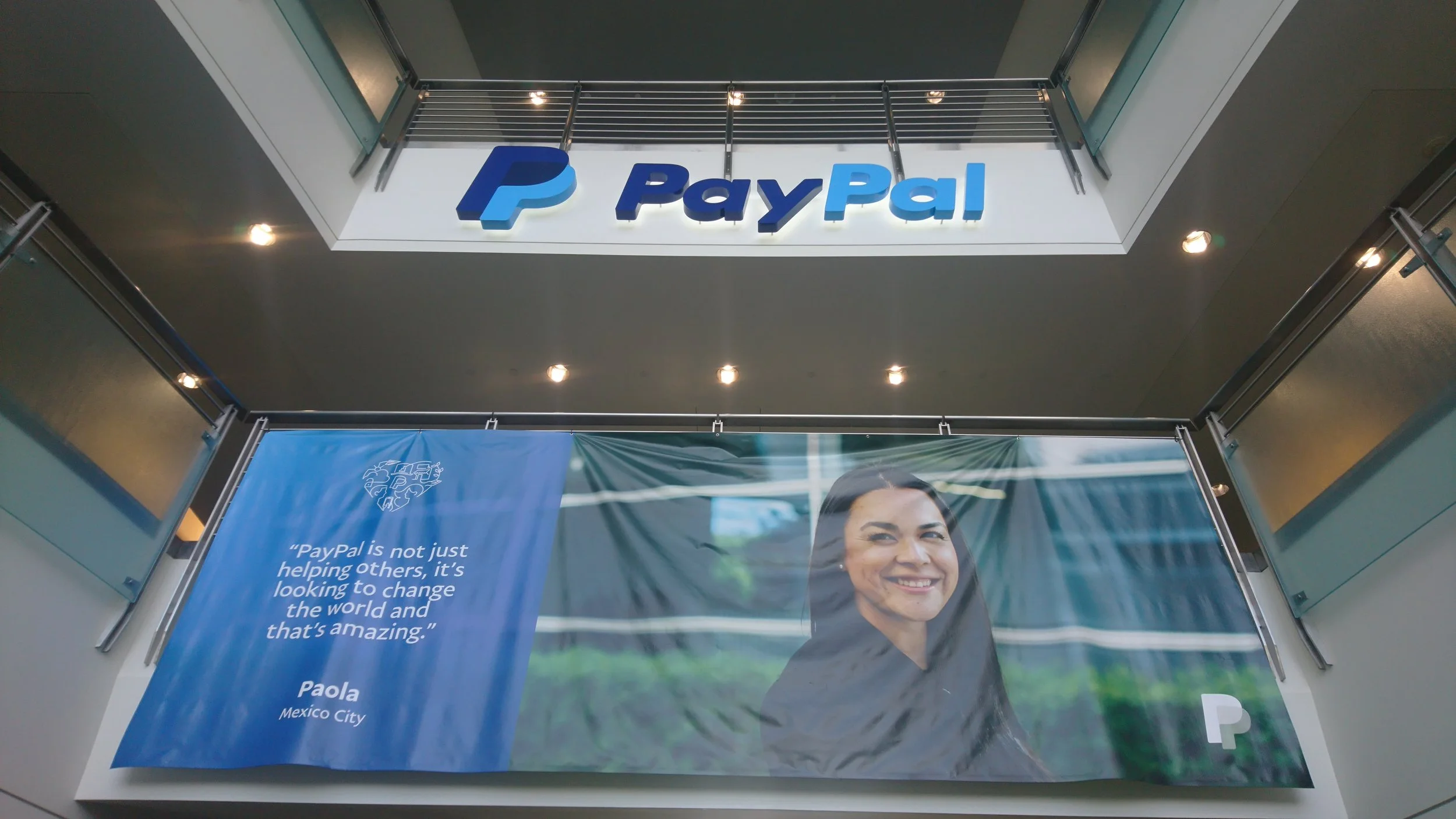Interior of a building with a large blue and white PayPal sign on the wall, and a banner featuring a smiling woman with dark hair and a quote from Paola from Mexico City.