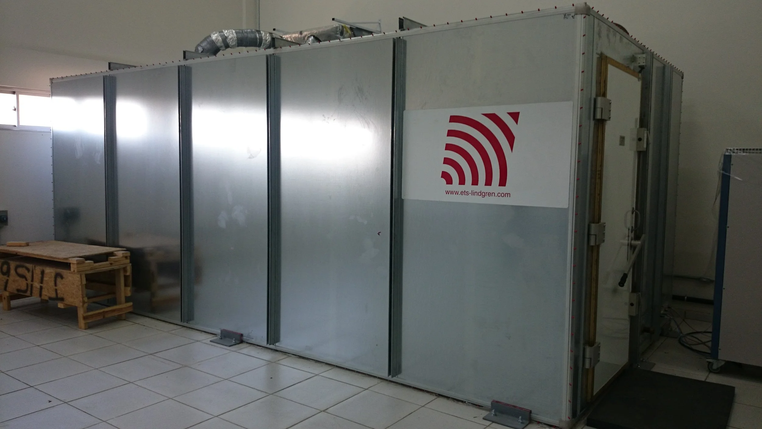 A large, industrial metal enclosure with a logo and website URL, situated on a tiled floor in an indoor setting.