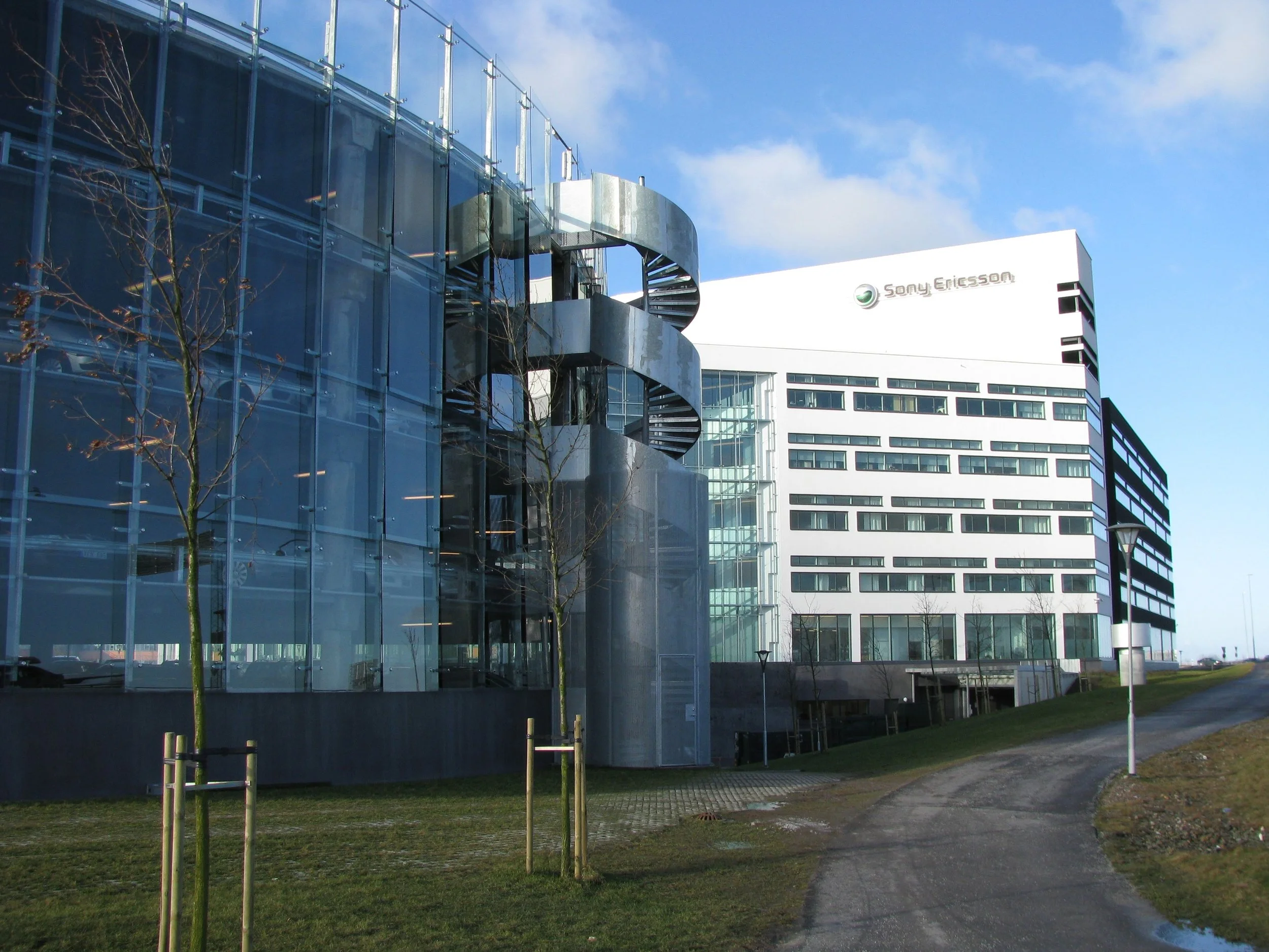 Modern office building with glass facade and the Sony Ericsson logo, with a curved metal staircase outside and a winding path leading towards it.