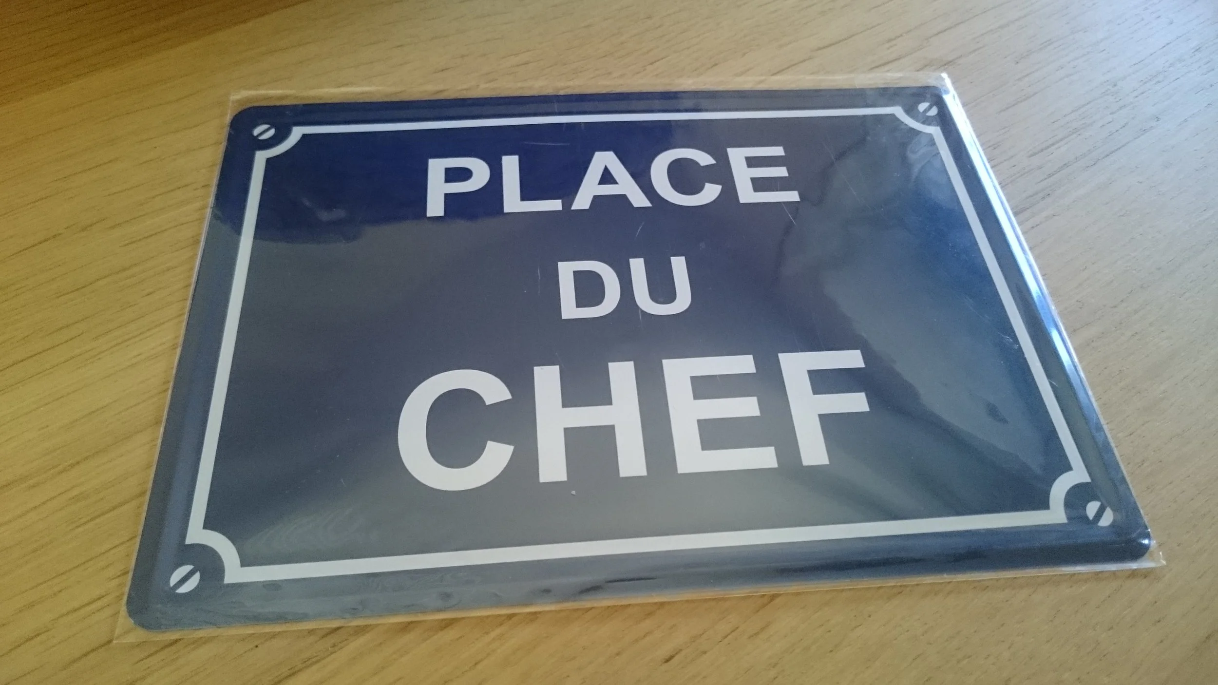 A metal sign with a blue background and white border that reads, 'Place du Chef,' placed on a wooden surface.