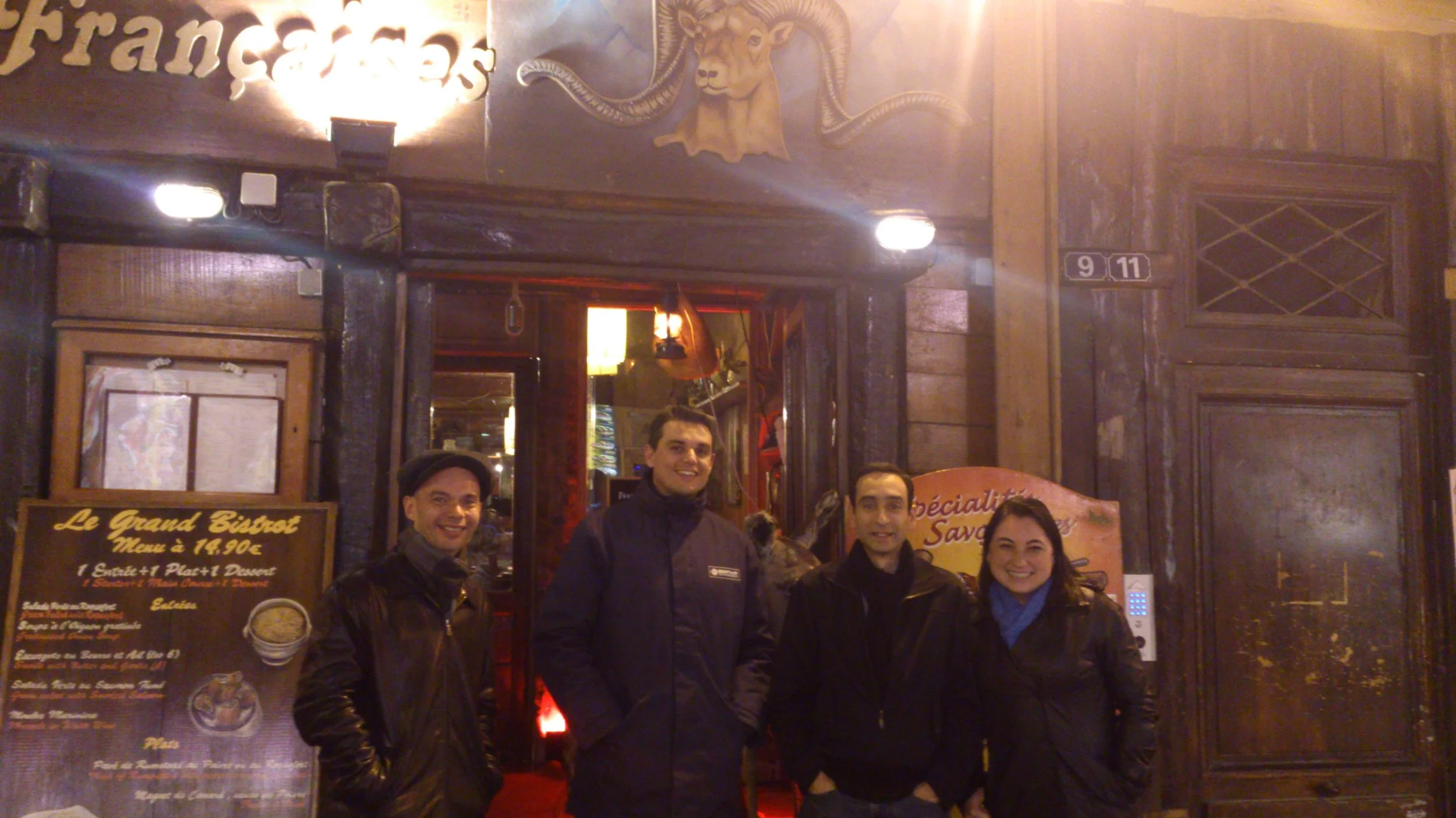 Four people standing in front of a restaurant entrance at night, smiling for a photo. The restaurant has a wooden exterior with a deer and ram head decoration above the doorway, and a menu board displaying prices and food options outside.