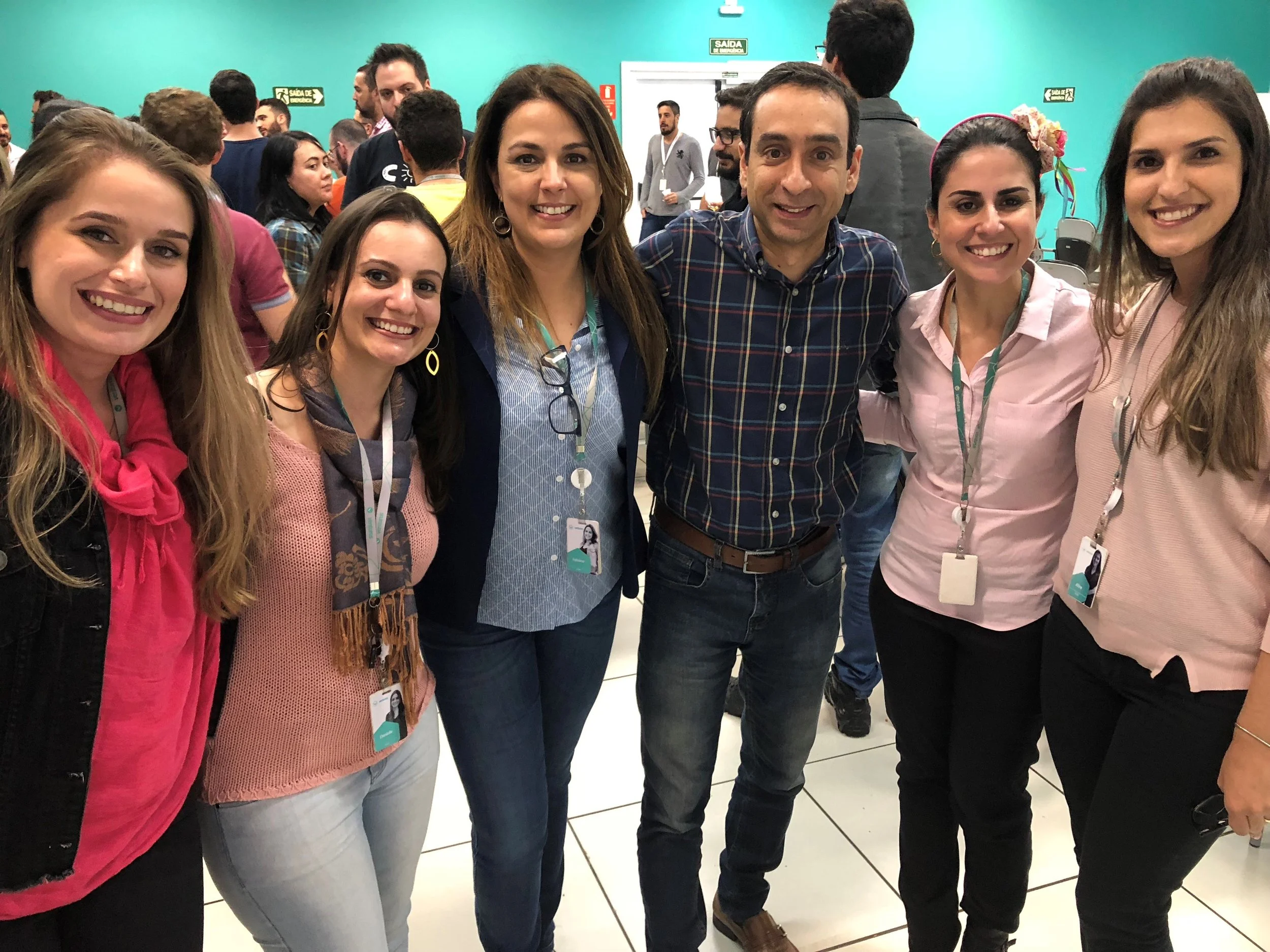 Group of seven smiling people, six women and one man, standing close together at an indoor event with teal walls and many people in the background.