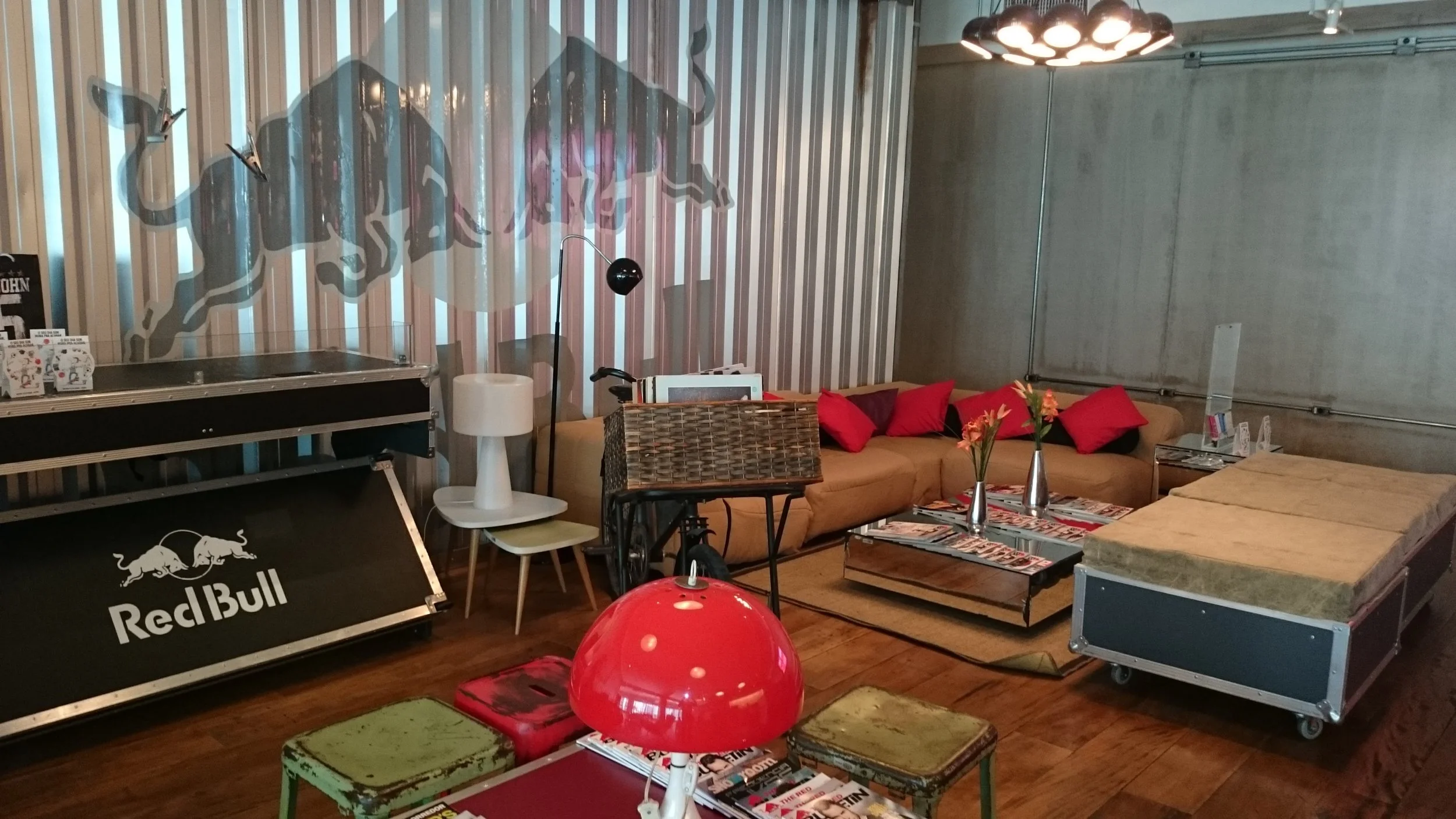 A cozy lounge with a Red Bull vending machine, a brown sofa with red pillows, a coffee table with magazines and vases, a green-stained stool, and a vintage-style red lamp, featuring a large mural of a bull on the wall.