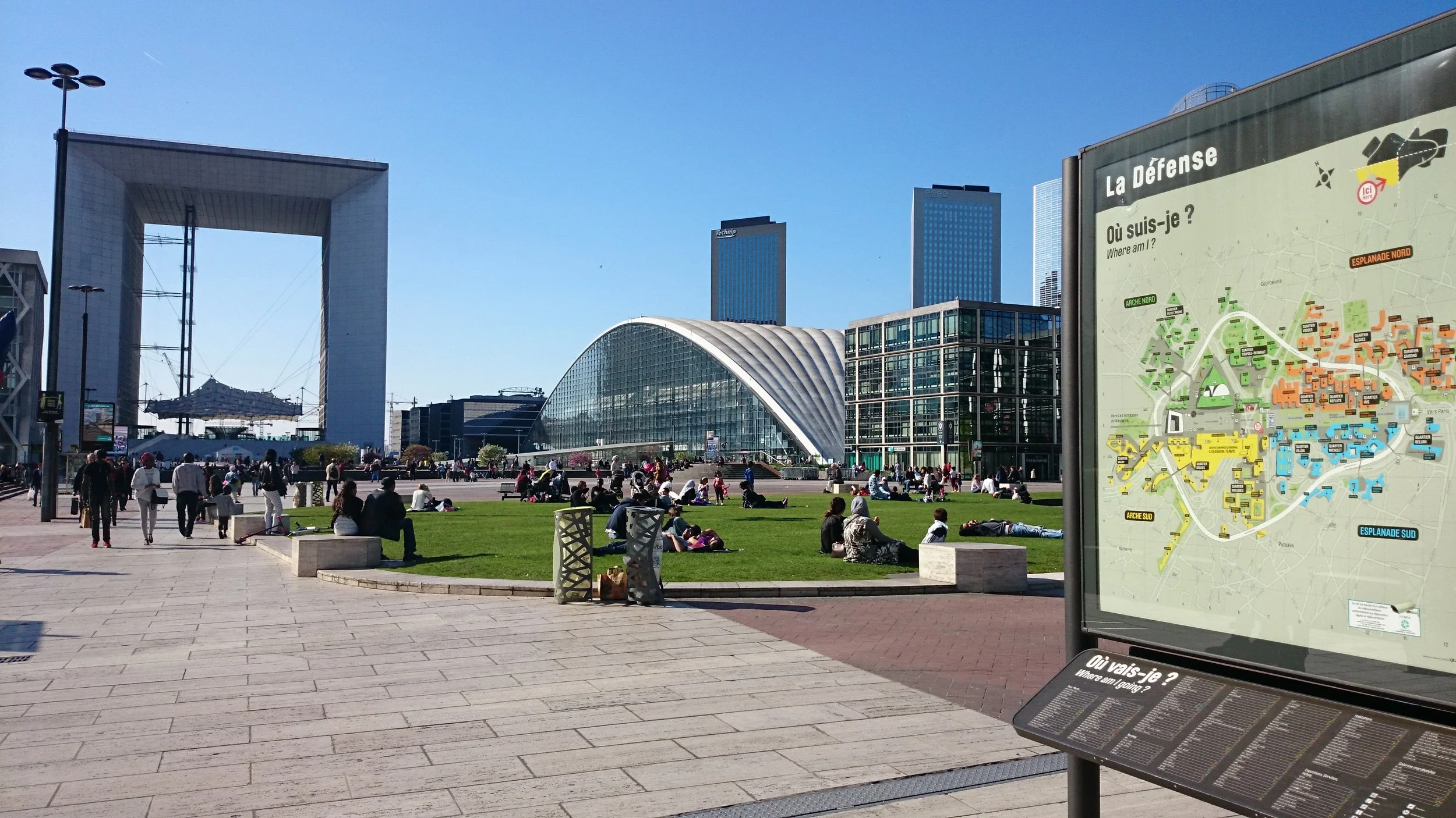 A busy public square with people sitting on the grass and walking on paved paths, modern buildings including a large arched glass structure and tall skyscrapers in the background, and a visible map kiosk on the right with a city map labeled 'La Défen
