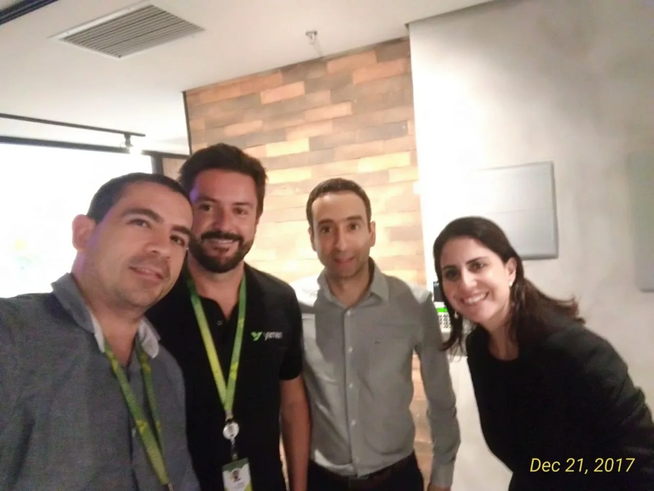 Four people standing close together indoors, smiling for a photo, with a wood-paneled accent wall behind them. It is December 21, 2017.