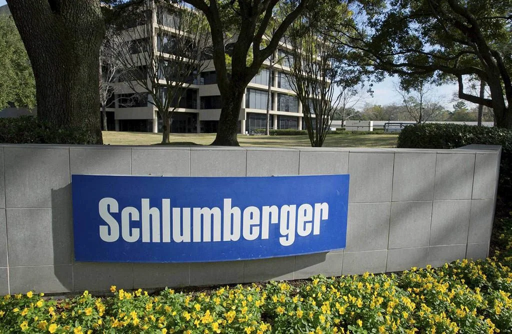 A large blue sign with the word 'Schlumberger' in white letters, set against a low gray wall with yellow flowers in front, trees, and a modern building in the background.