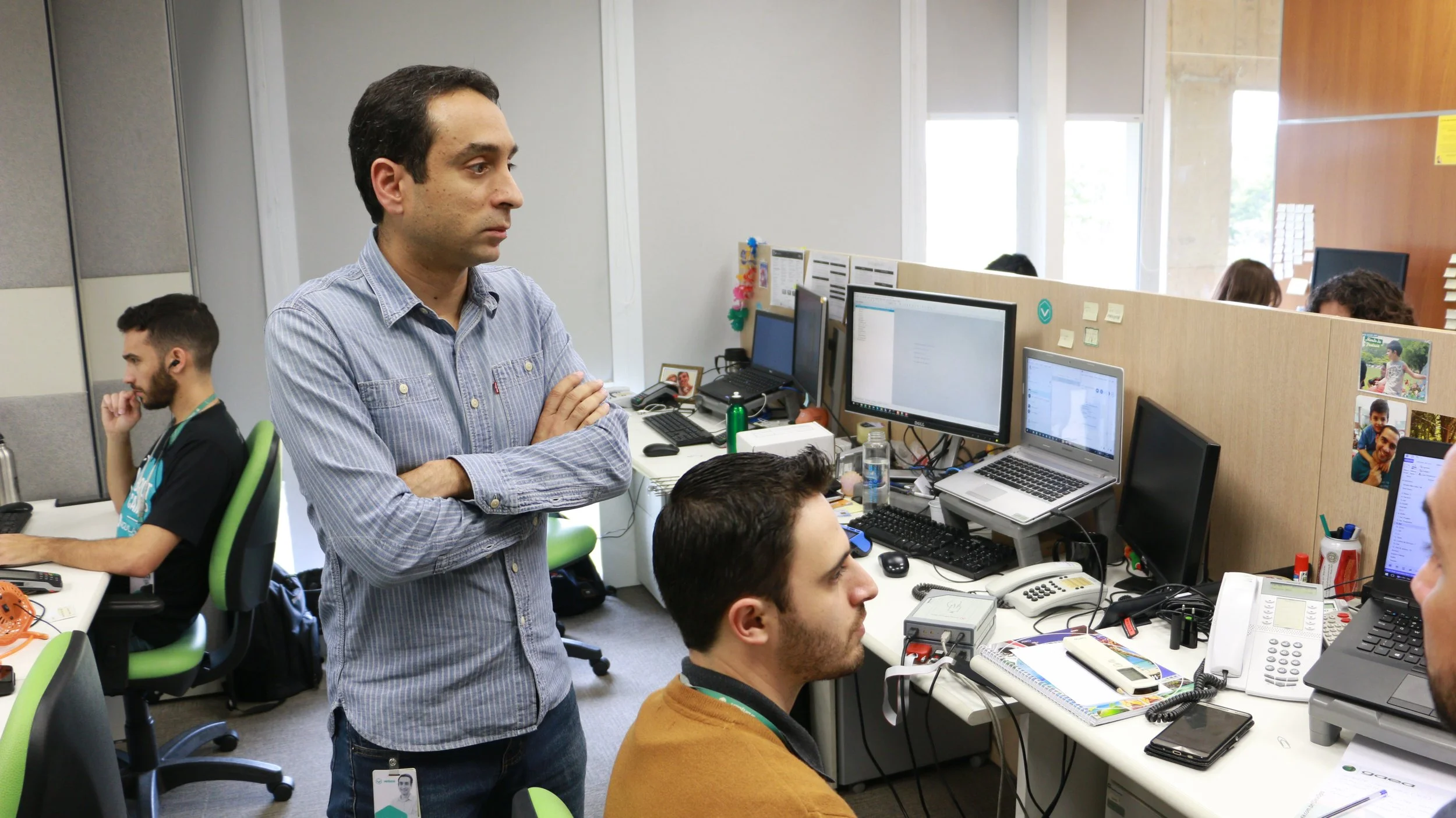 Office scene with multiple employees working at their desks, a man standing with crossed arms observing, and several computer monitors and office supplies on the desks.