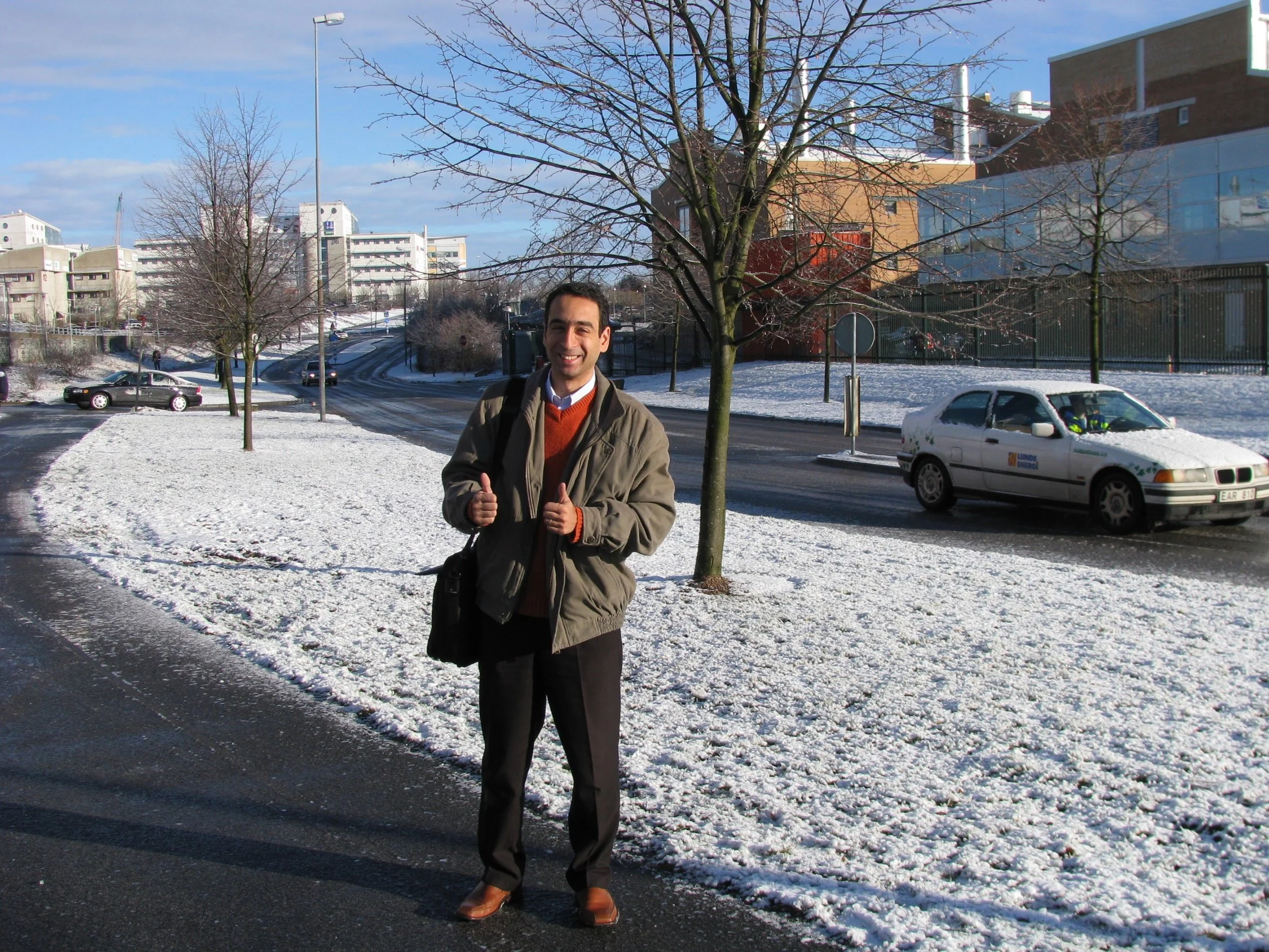 A man standing on a snowy street giving two thumbs up. He is wearing a beige jacket, red sweater, and black pants, with a black shoulder bag. There are leafless trees and cars on the road, with buildings in the background under a clear blue sky.