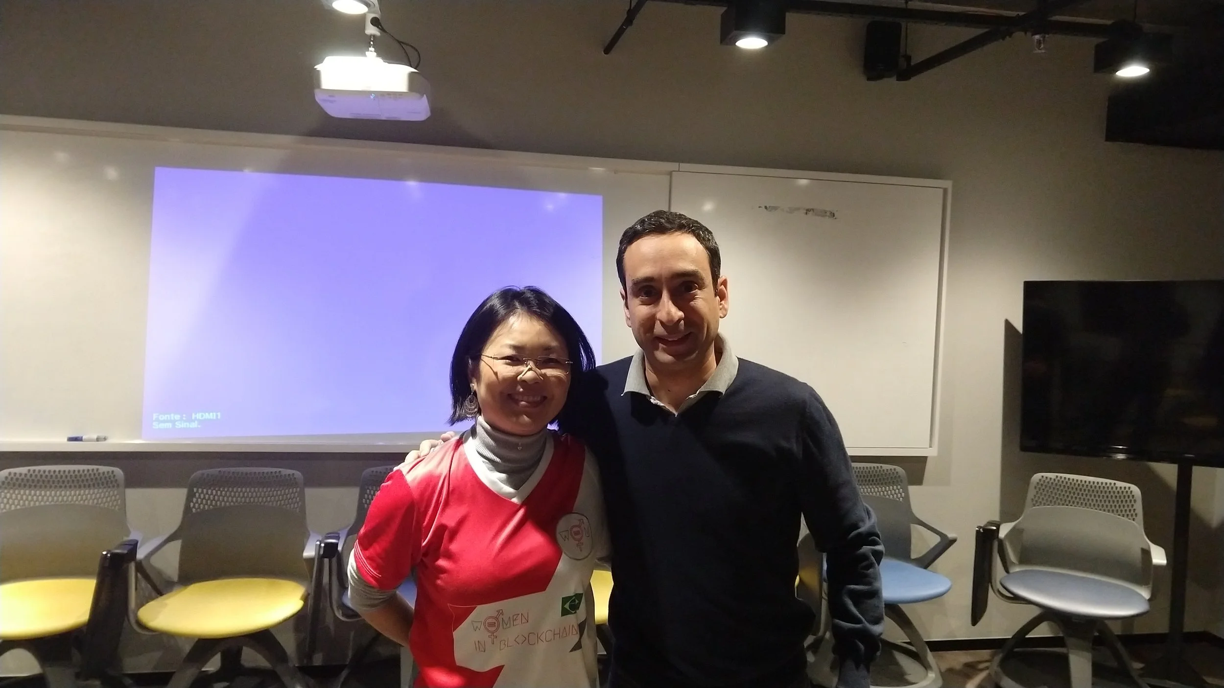 A woman wearing glasses and a red and white T-shirt stands next to a man in a black sweater. They are smiling and standing close together in a room with chairs, a projection screen, and a whiteboard.
