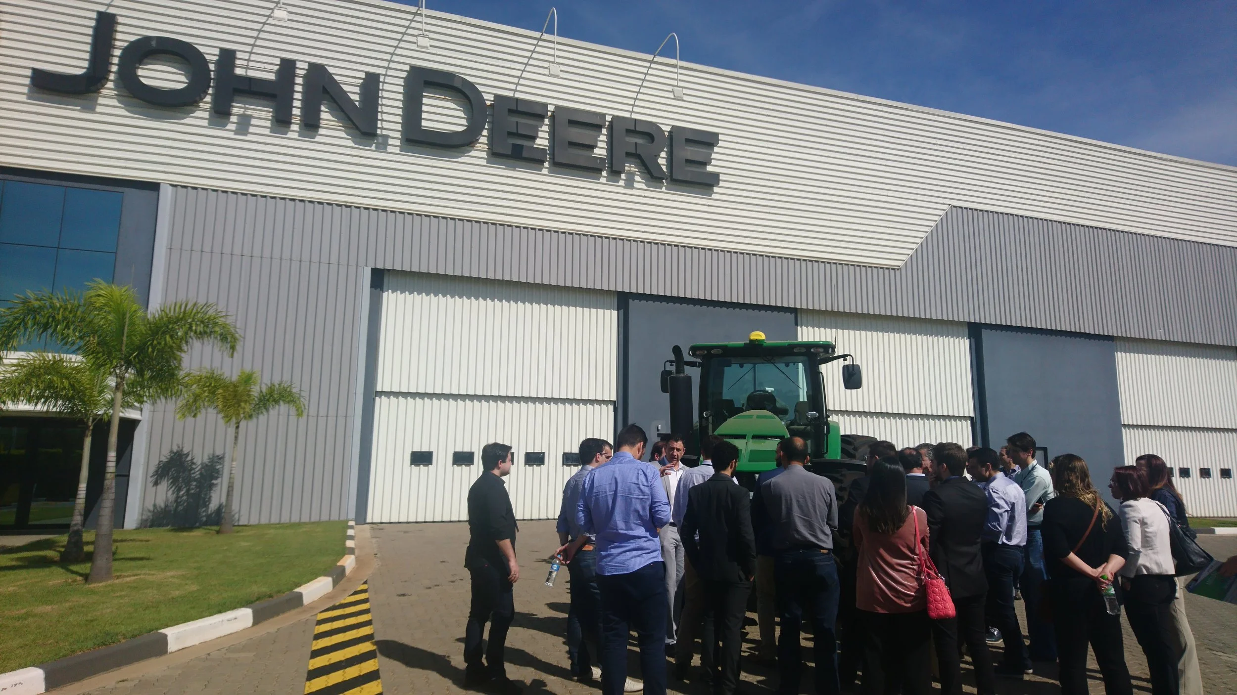 A group of people standing outside a large building with the name 'JOHN DEERE' on it, gathered around a green tractor for a presentation or demonstration.