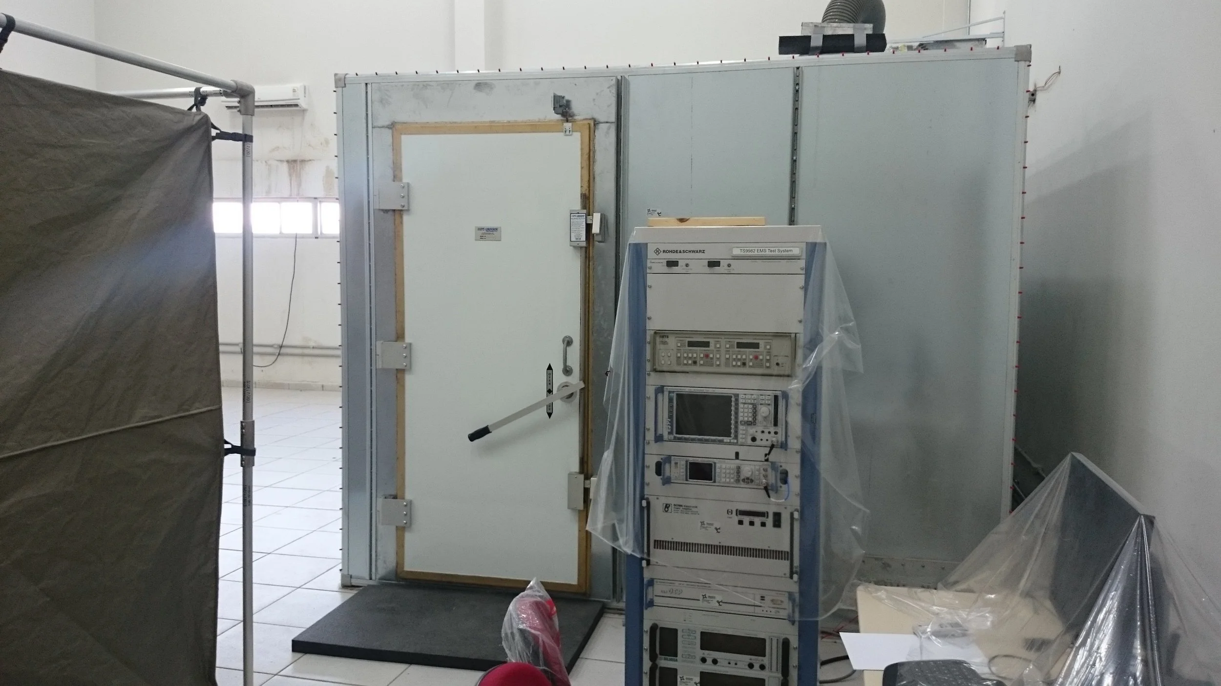A room with a white door and a rack of electronic testing equipment, with some plastic covering parts of the equipment.