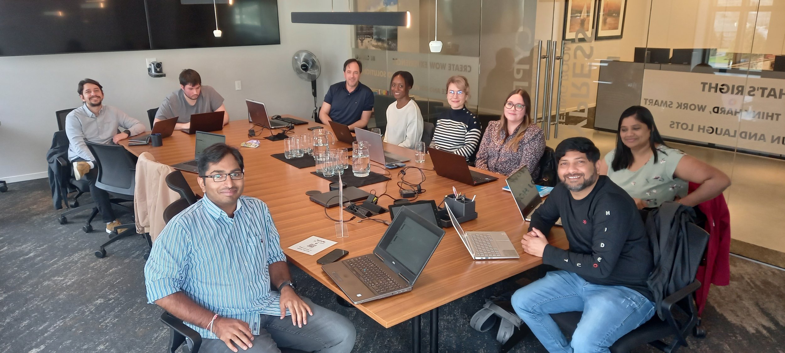 Group of ten diverse professionals sitting around a large conference table with laptops and glass pitchers in a modern office conference room.