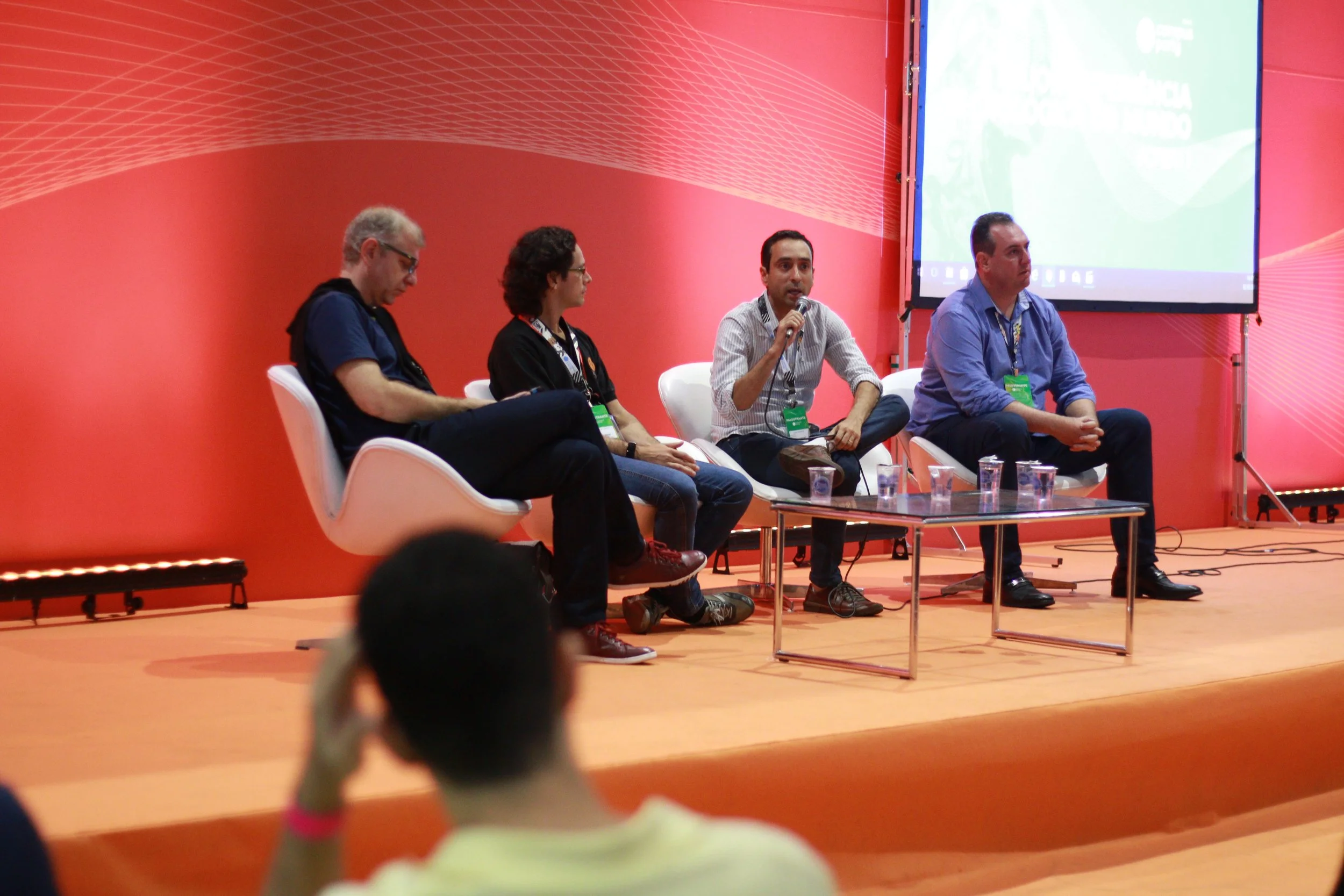 Four people sitting on a stage in front of a red background, participating in a panel discussion at a conference. One person is speaking into a microphone, while the others listen. There are four glasses of water on a small table in front of them.