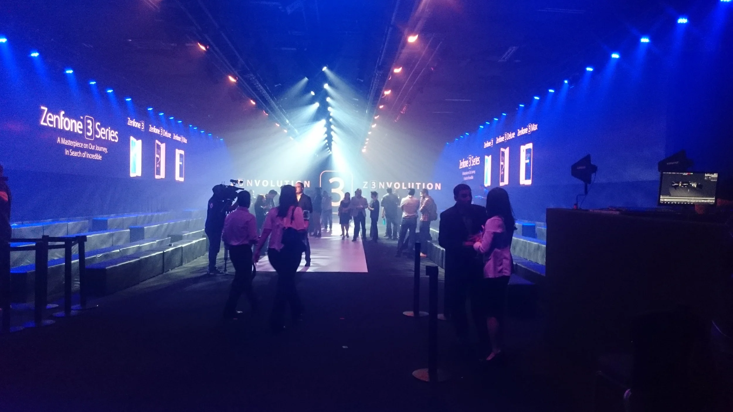 An indoor event space with blue and purple lighting, people mingling, and large screens displaying promotional content for the Zenfone 3 Series smartphones.