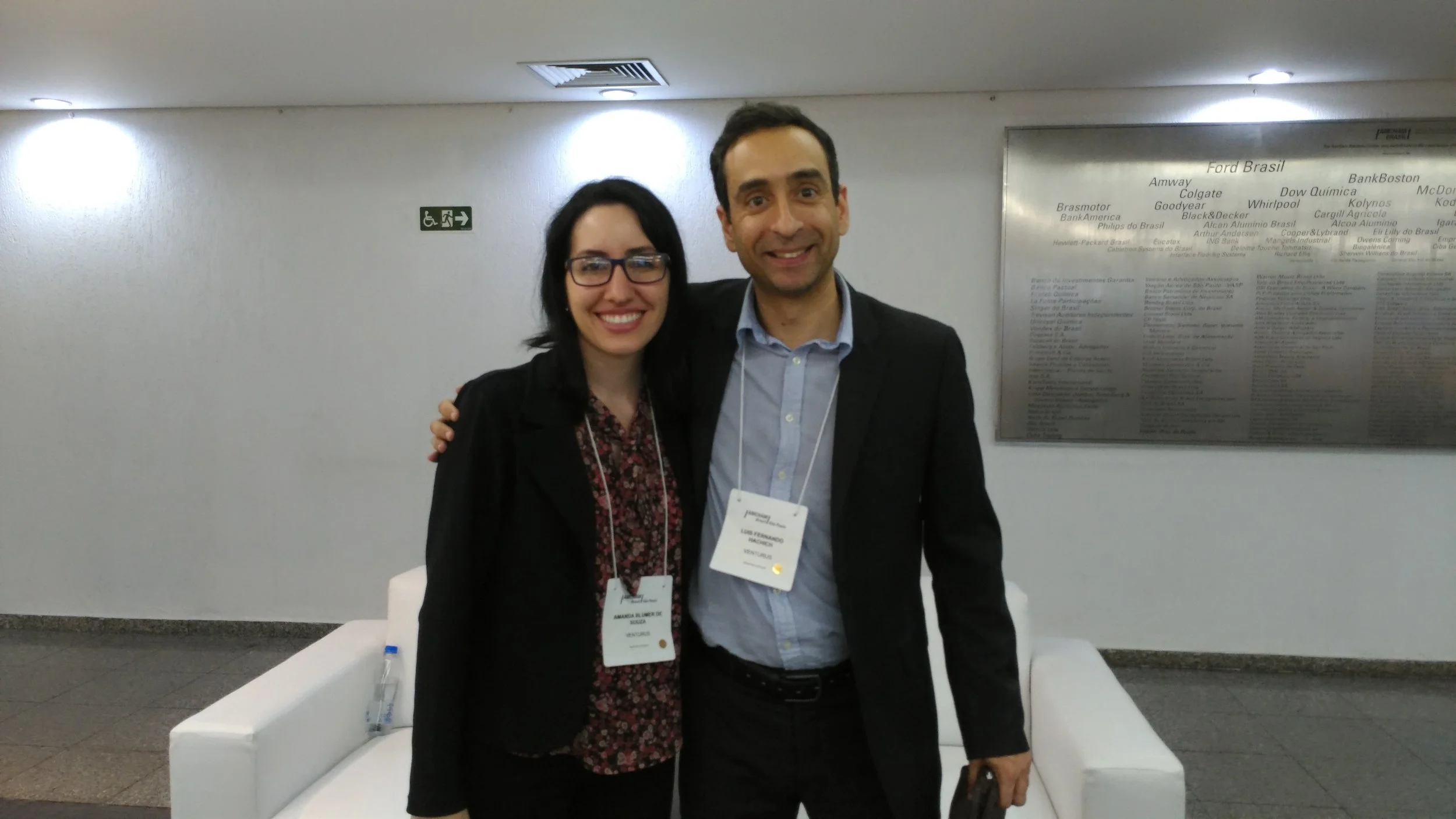 Two people, a woman with glasses and a man, standing together indoors, smiling, with conference badges around their necks.