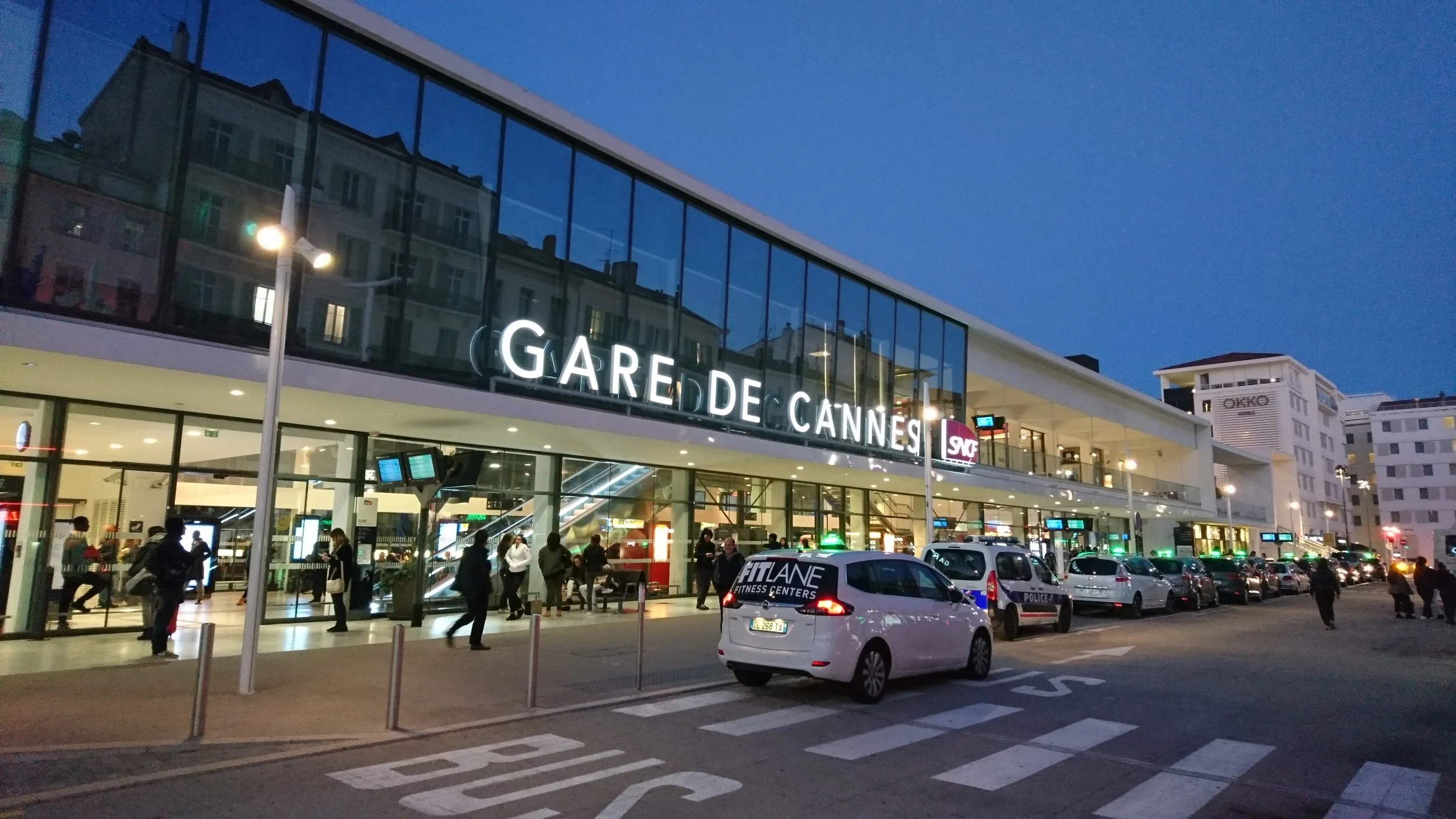 Exterior view of Gare de Cannes train station at dusk, with people walking outside and parked cars along the street.