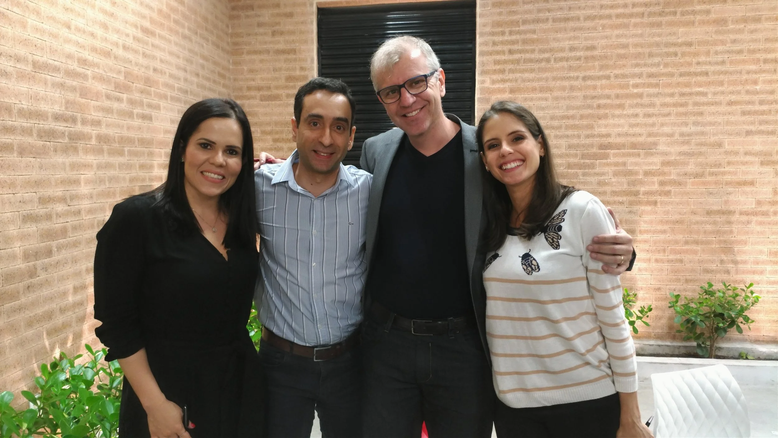 Four people standing together, smiling, in an indoor setting with brick walls and some green plants in the background.