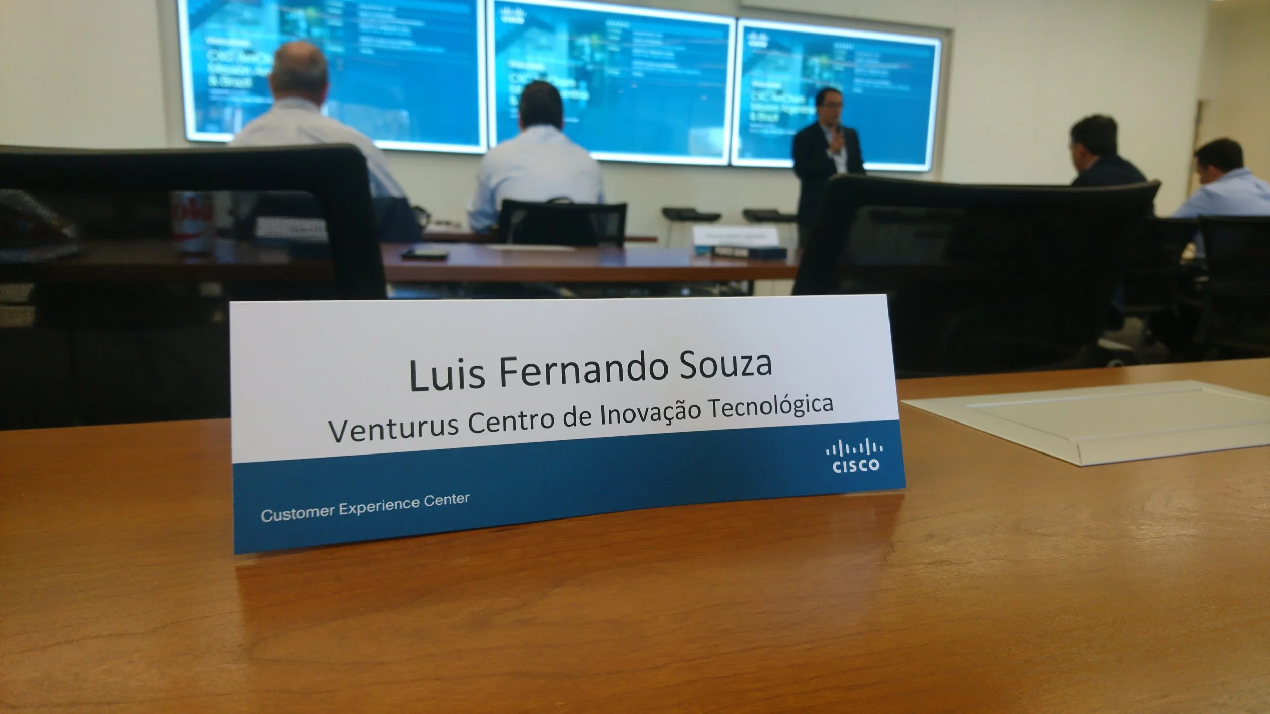 Nameplate on a desk in a conference room showing 'Luis Fernando Souza, Venturus Centro de Inovação Tecnológica, Customer Experience Center, Cisco'. In the background, people are sitting at tables with large screens on the wall.